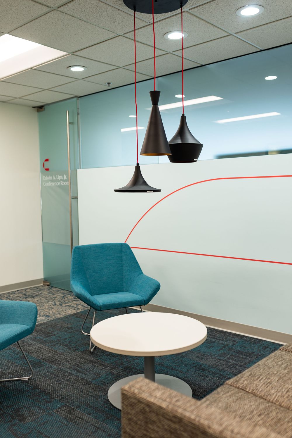Modern office lounge with teal chairs, white round table, and black pendant lights hanging from red cords near a glass conference room.