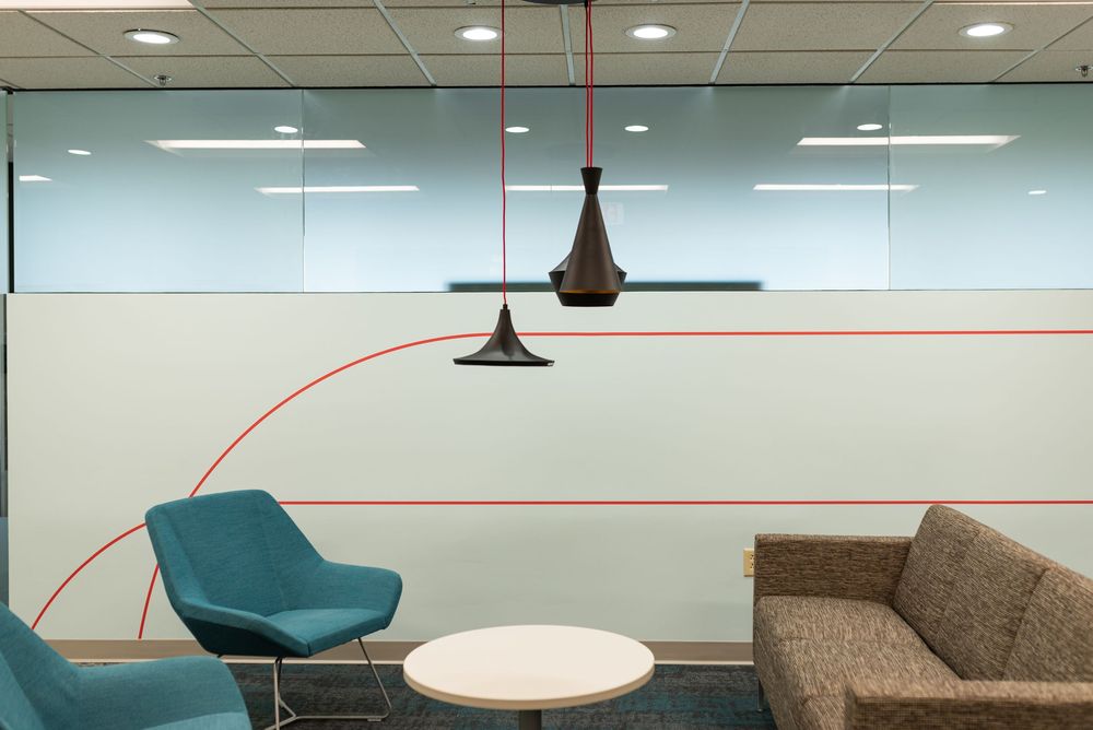 Modern office lounge with teal chairs, brown sofa, round table, and stylish black pendant lights near frosted glass wall.