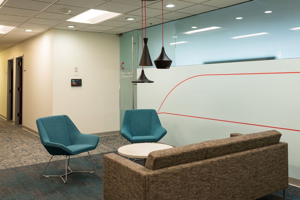 Modern office lounge with teal chairs, pendant lights, glass partition with red design, and neutral sofa seating area.