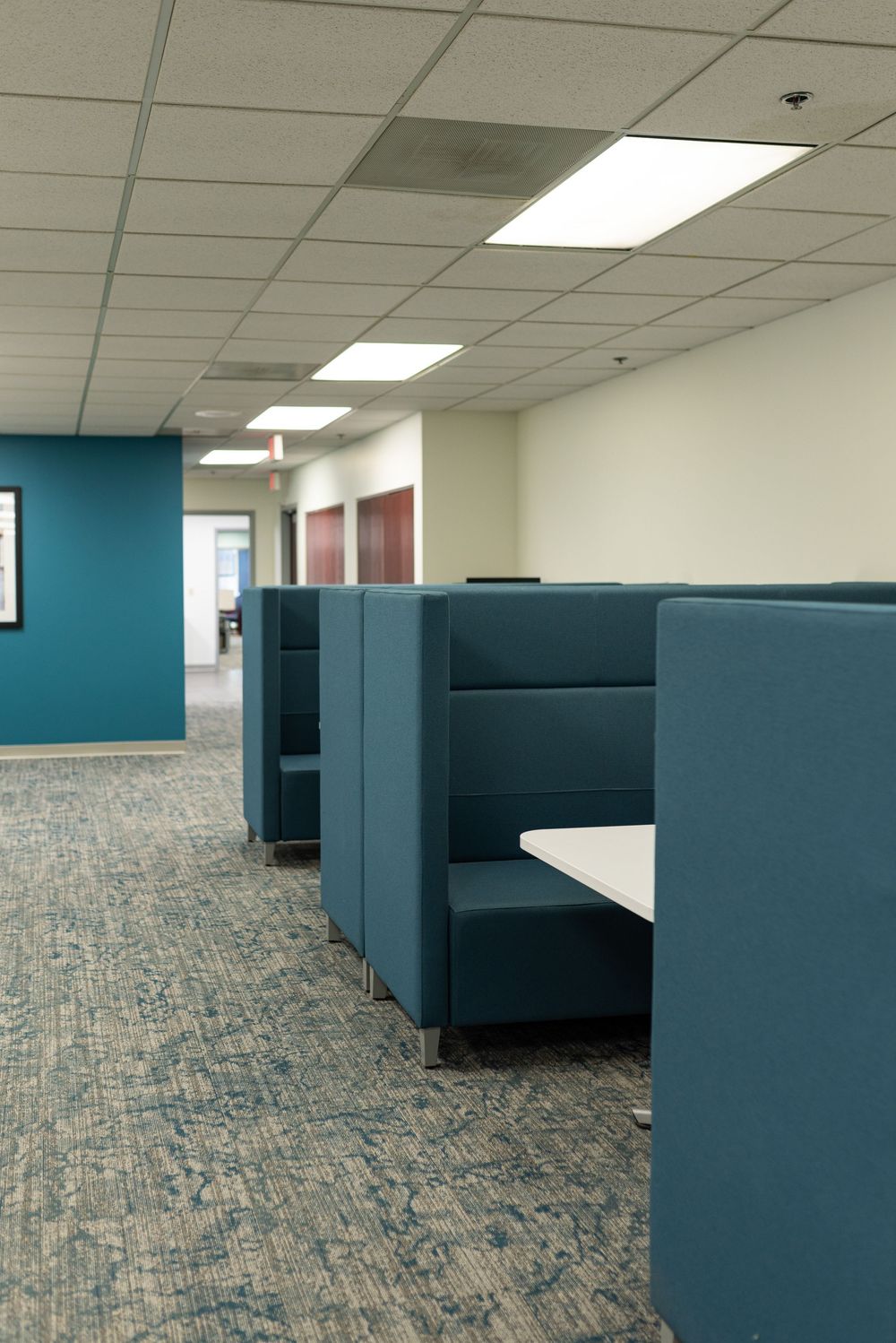Modern office space featuring teal upholstered booth seating cubicles with white tables on patterned carpet flooring.