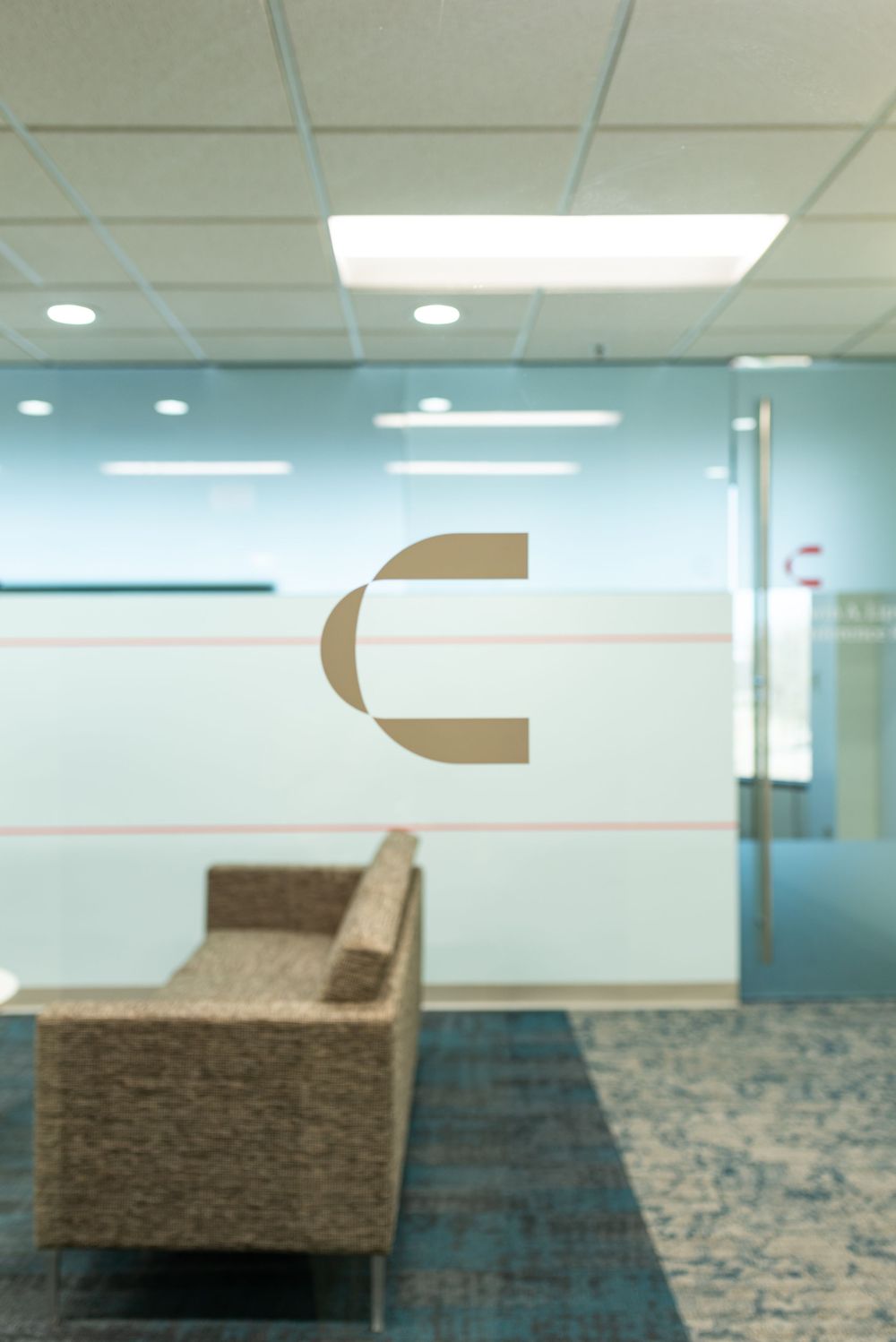 Modern office lobby with blue glass partitions, a gold letter C logo, woven armchair, and patterned carpet flooring.