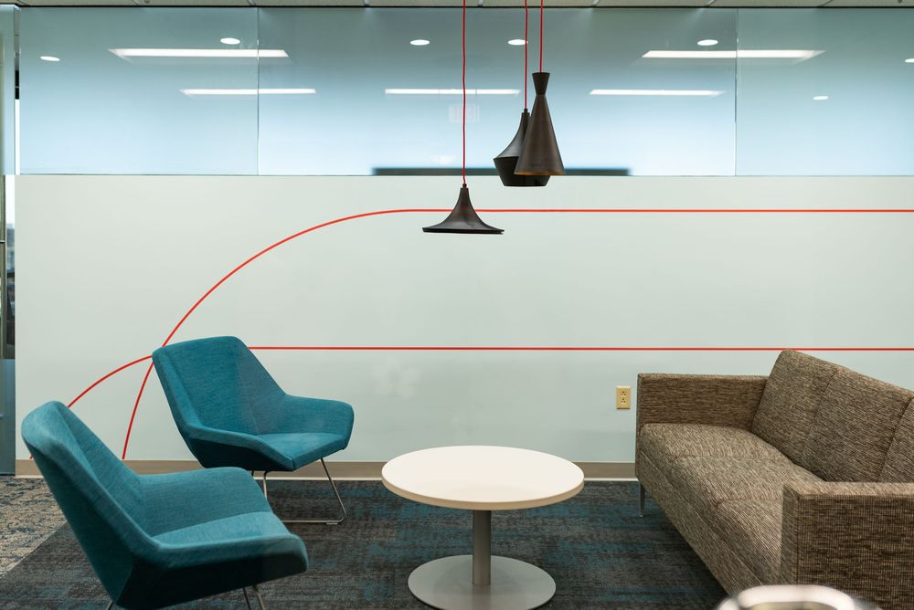 Modern office lounge with teal chairs, gray sofa, round white table, pendant lights, and red line decor on glass wall.
