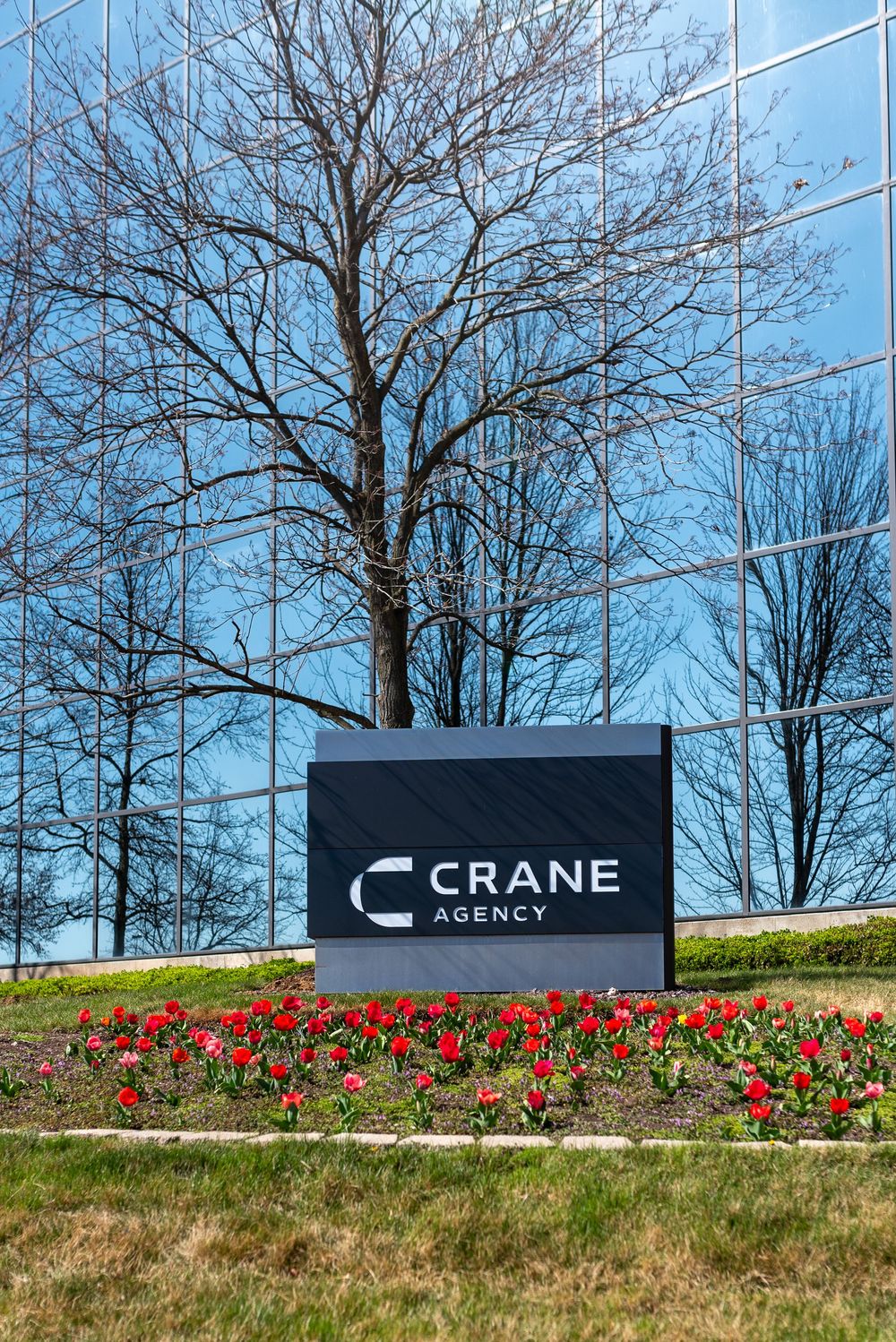 Crane Agency sign in front of a glass building with red tulips and bare trees reflecting in the mirrored facade.