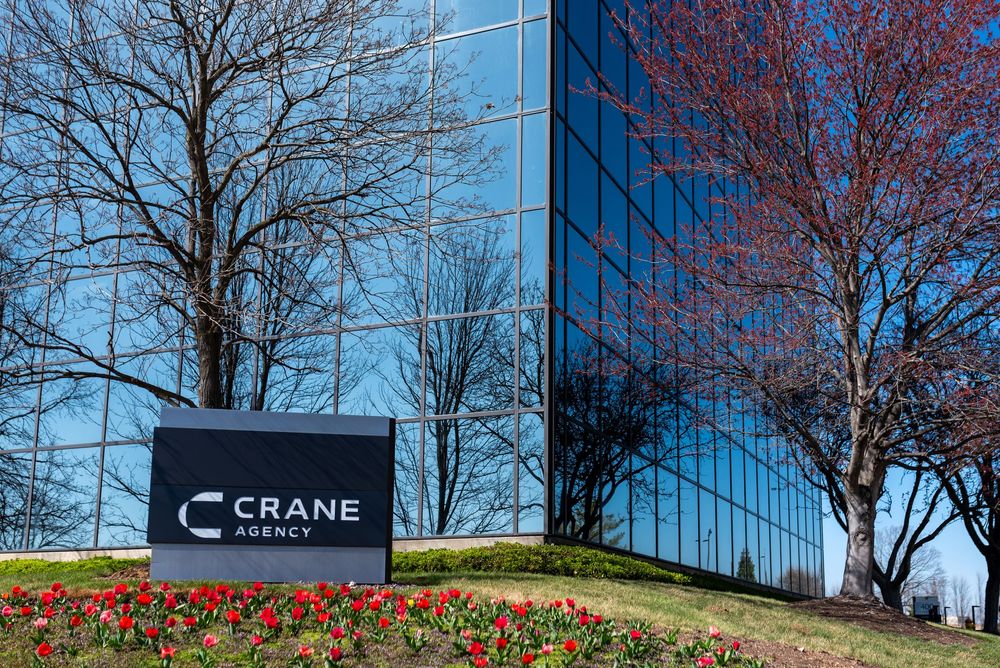 Crane Agency office building sign with tulips and bare trees in front of a modern glass corporate building.