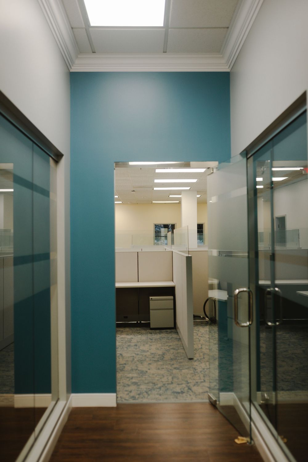 Modern office hallway with teal accent wall and glass partition doors leading to cubicles.