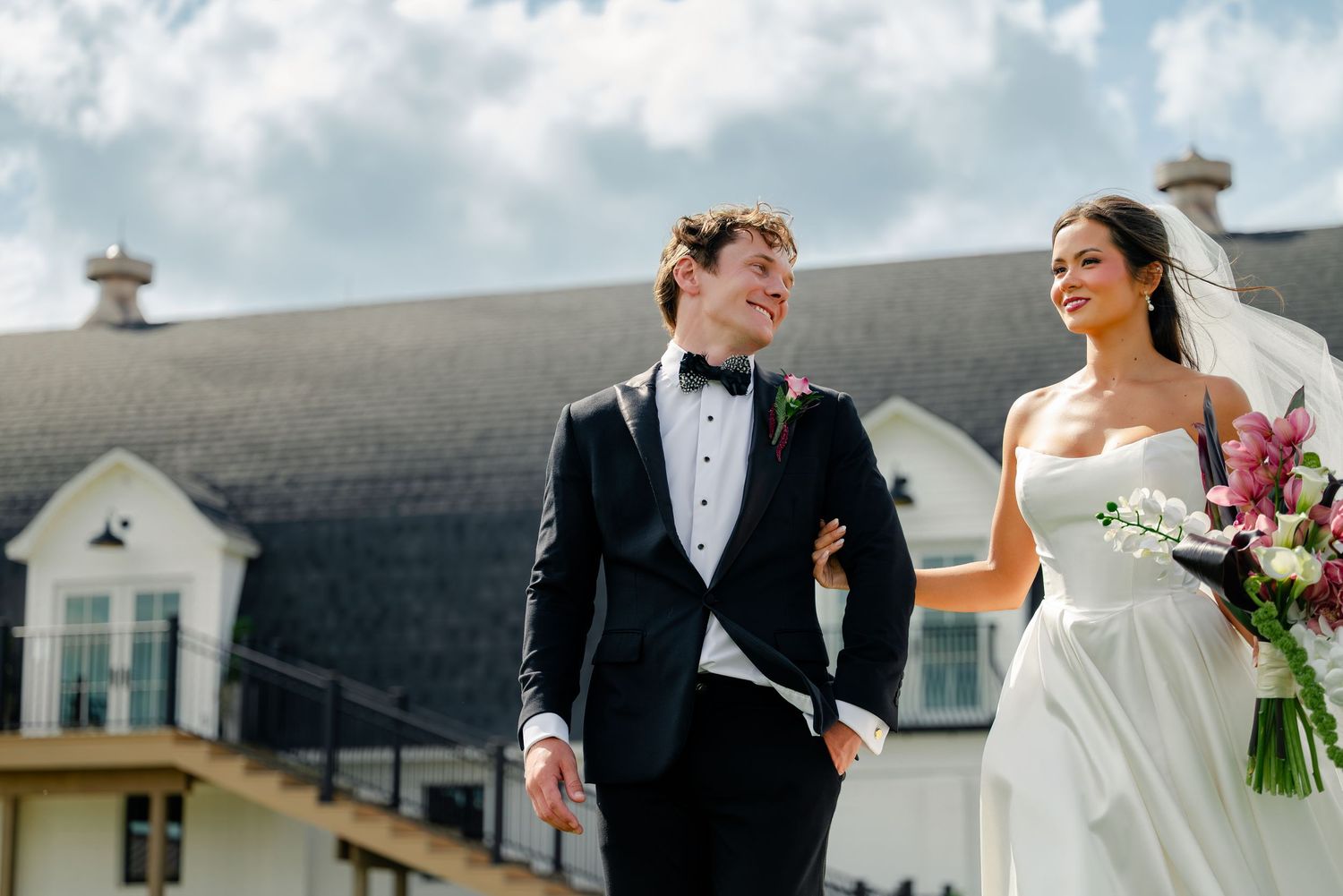 A bride and groom walk arm in arm outdoors, smiling, with the bride holding a pink floral bouquet and wearing a white gown.