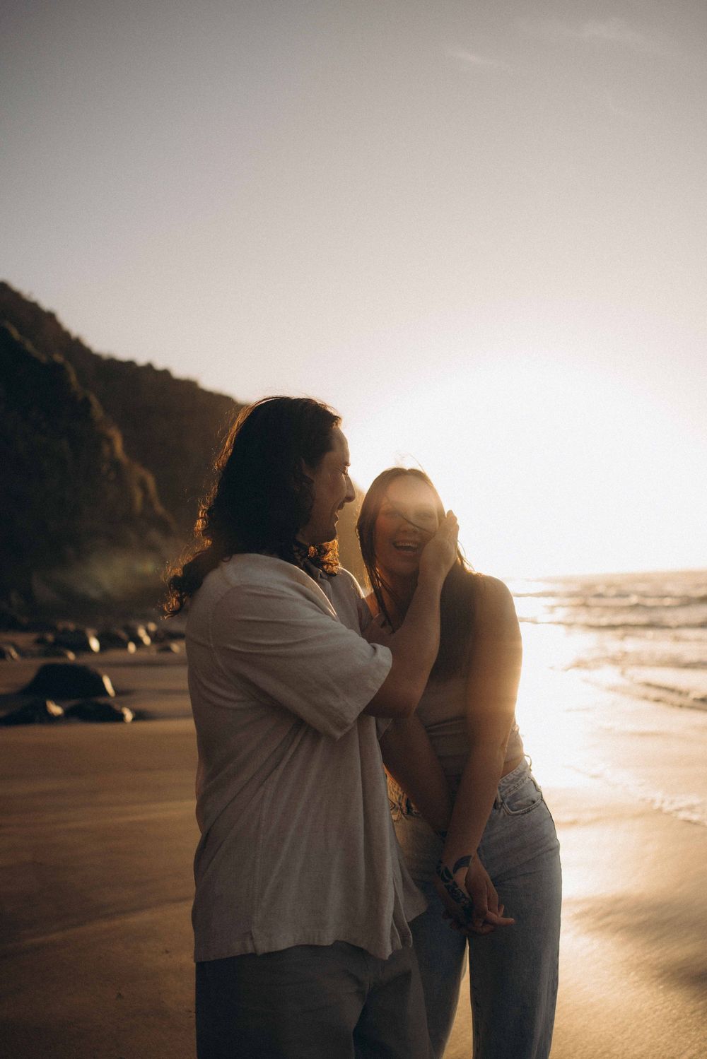 Couple laughing together on a sunlit beach at golden hour with ocean waves and rocky cliffs in the background.