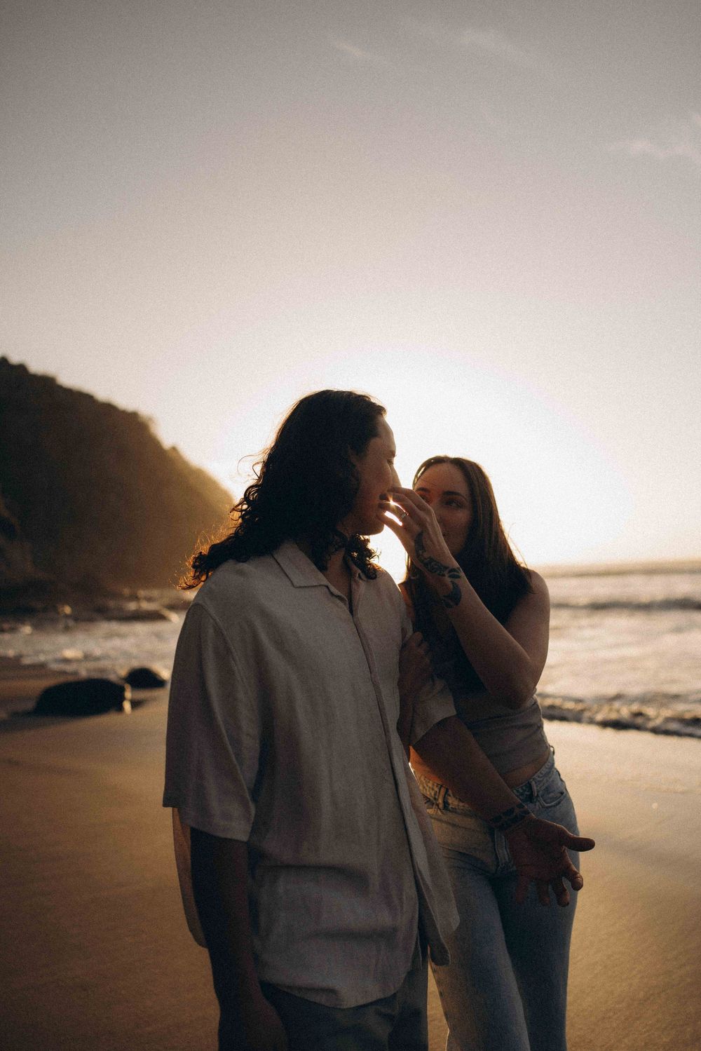 Couple sharing a tender moment on a sandy beach at golden sunset with ocean waves and rocky cliffs.