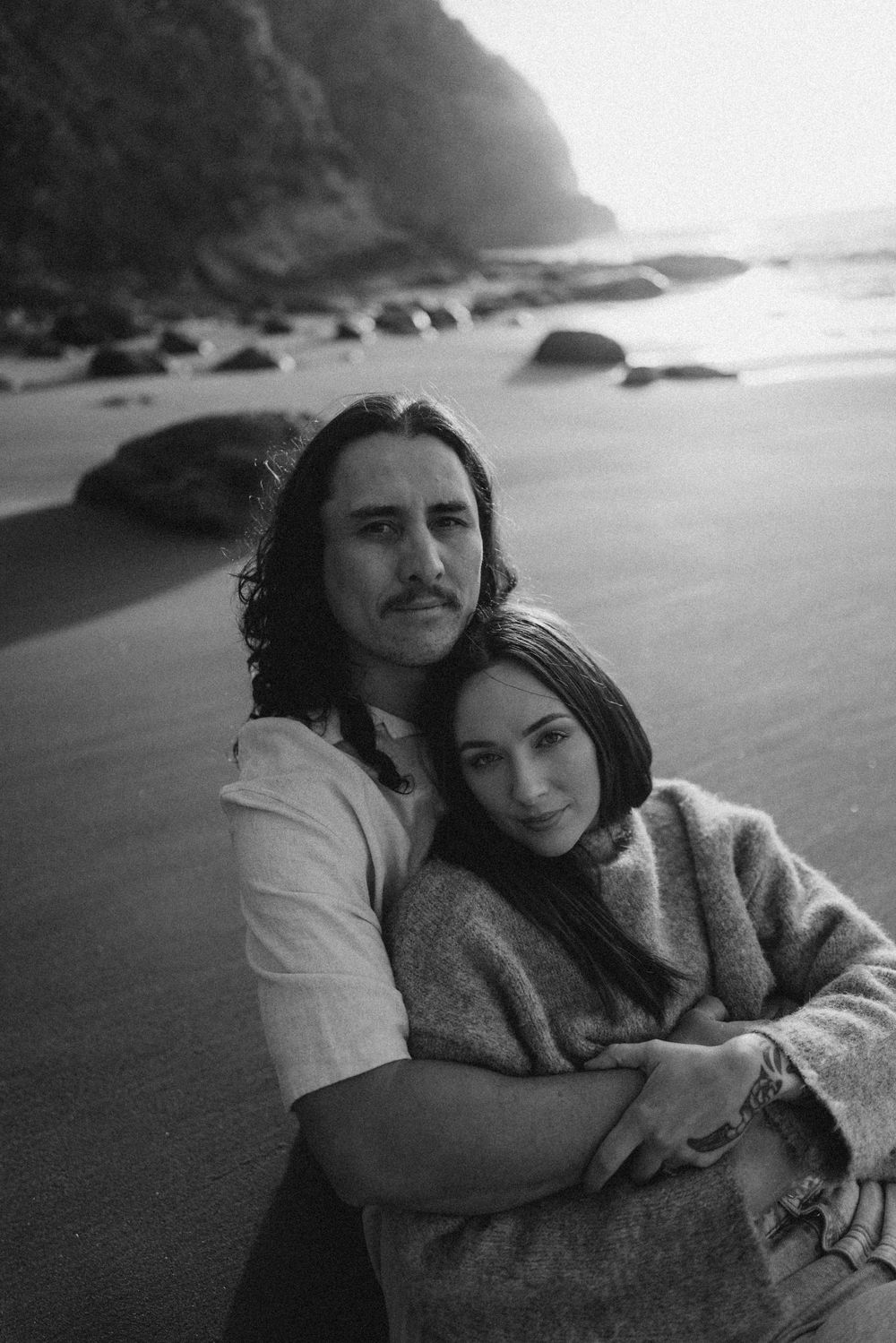 Black and white photo of a couple embracing on a rocky beach, with ocean waves and cliffs in the background.