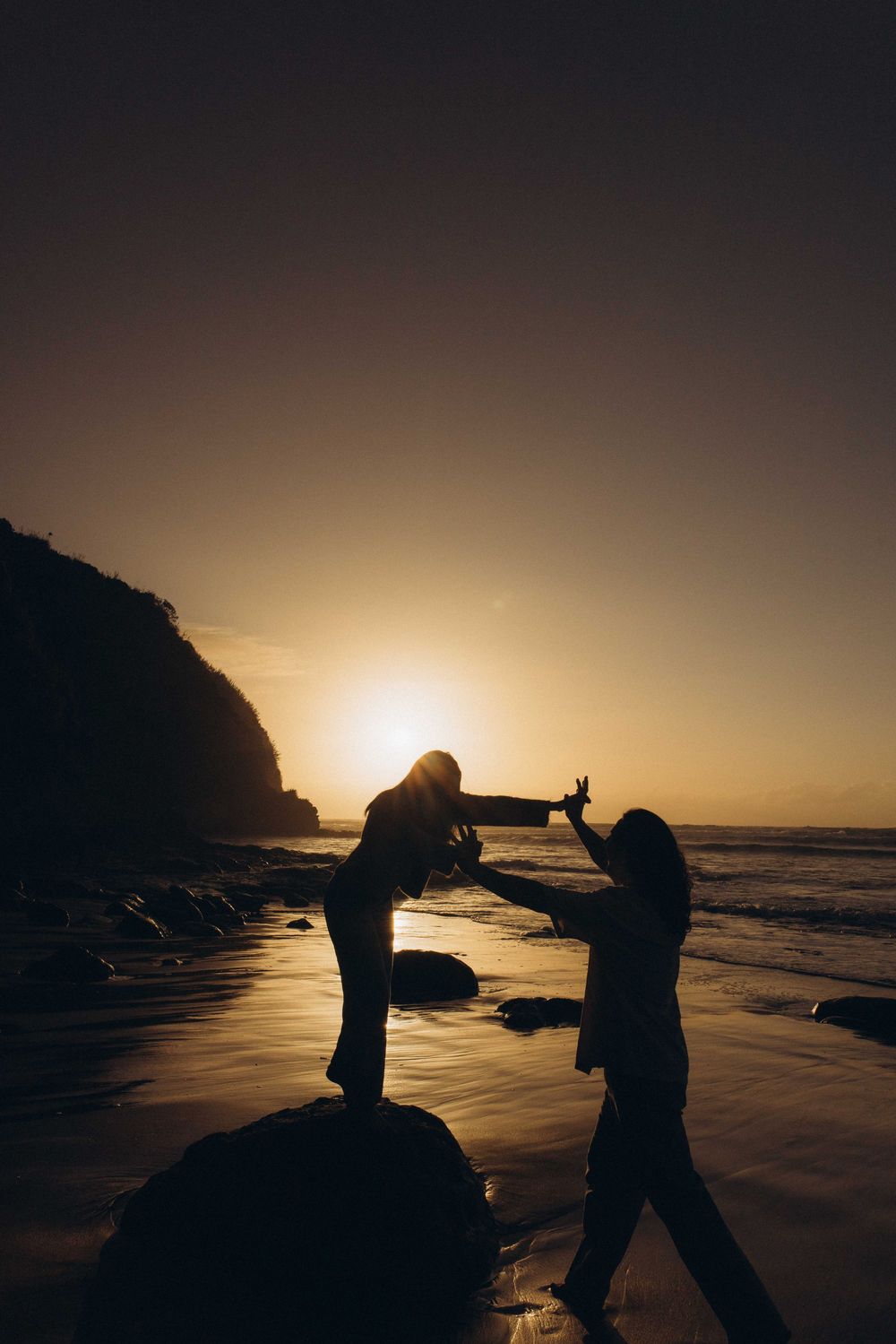 Two silhouettes dance on a beach at golden sunset with ocean waves and rocky cliffs in the background.