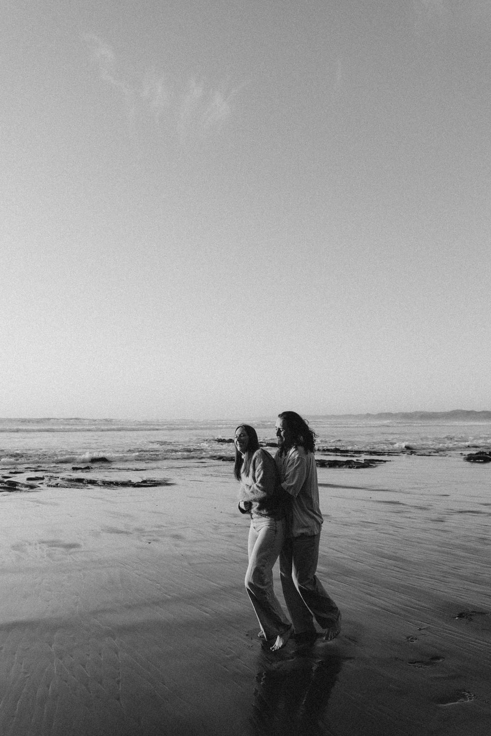 Two people embrace on a reflective beach shore in a black and white photo with ocean waves and clear sky.