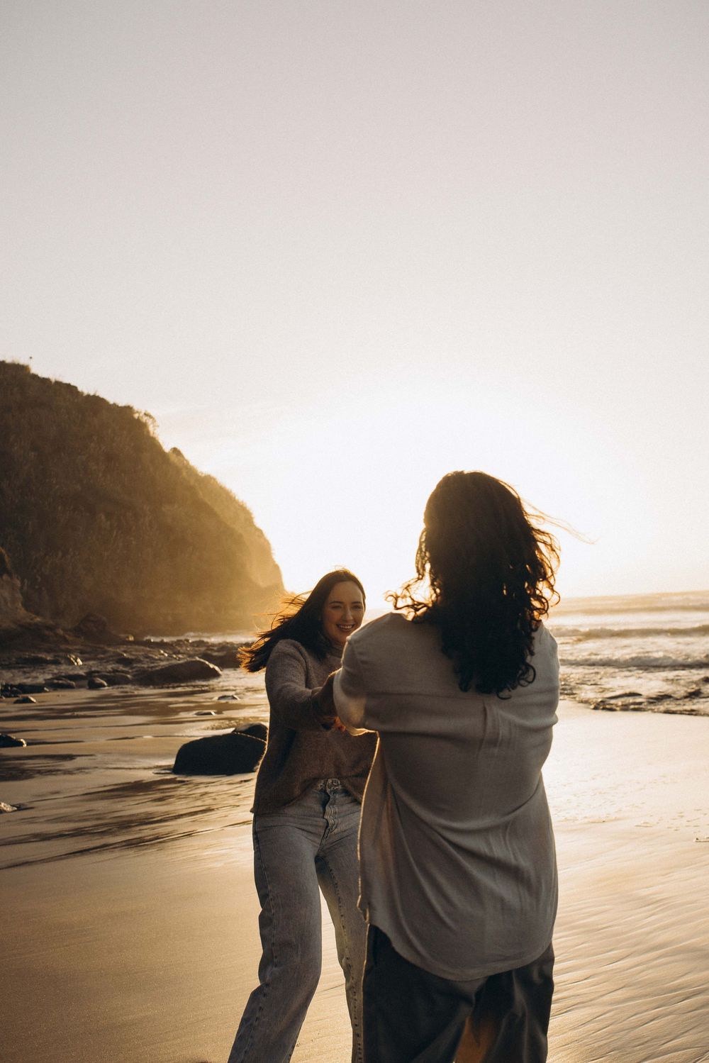 Two women dancing and laughing together on a sunlit beach at golden hour with rocky cliffs in the background.