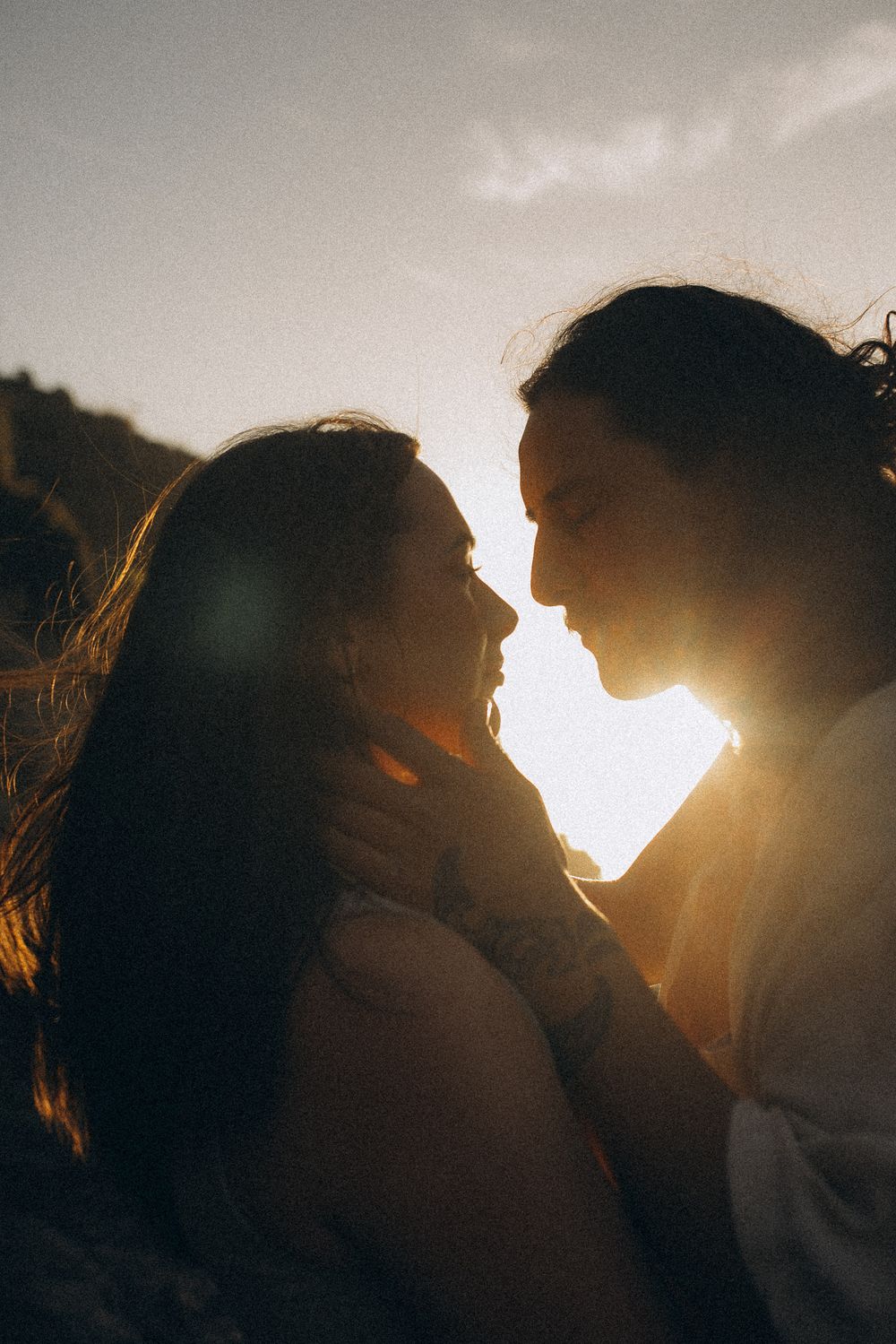 Romantic couple sharing an intimate moment at golden sunset, backlit silhouette with warm sun flare between them outdoors.