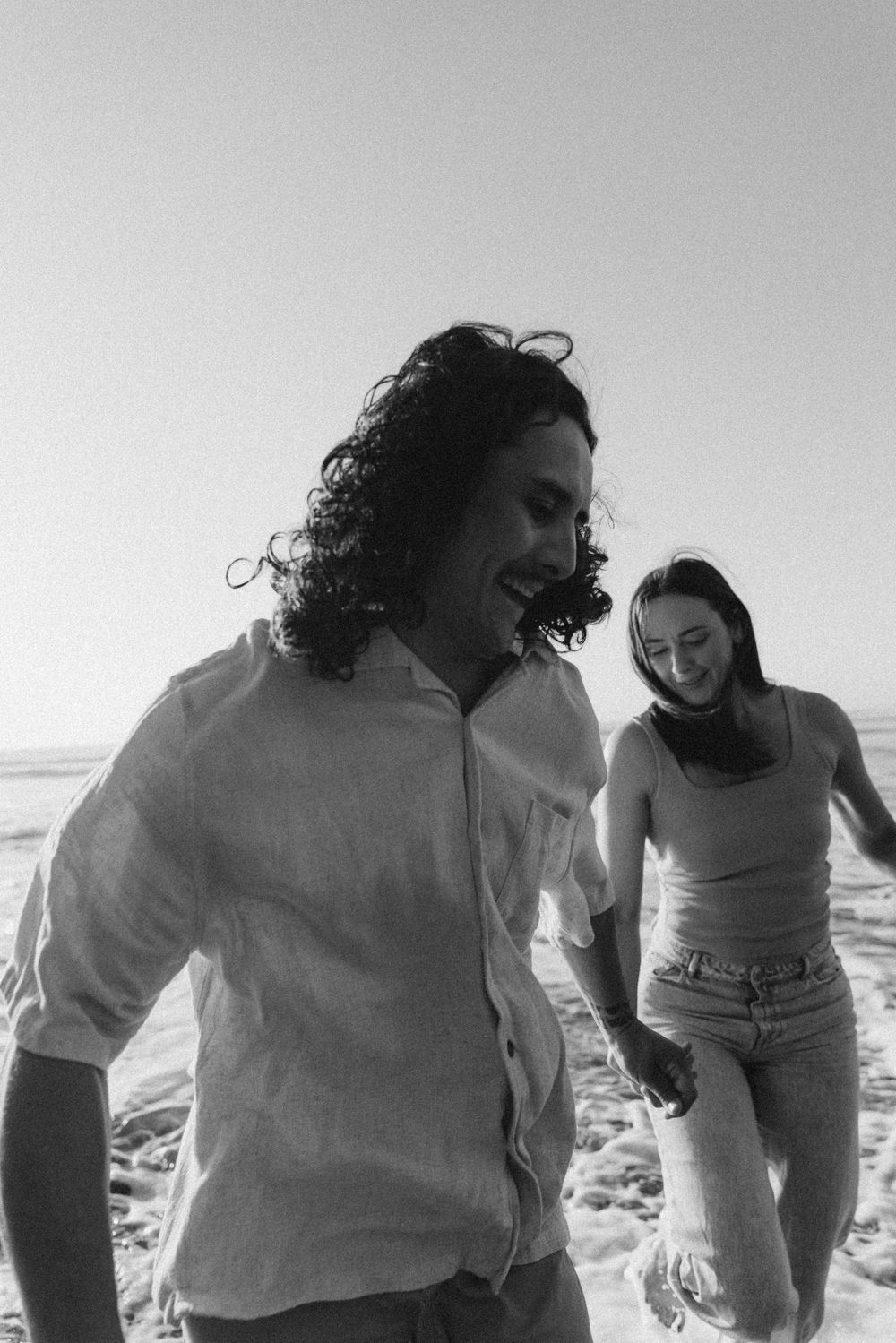 Black and white photo of a laughing couple holding hands and running through ocean waves on a sunny beach.