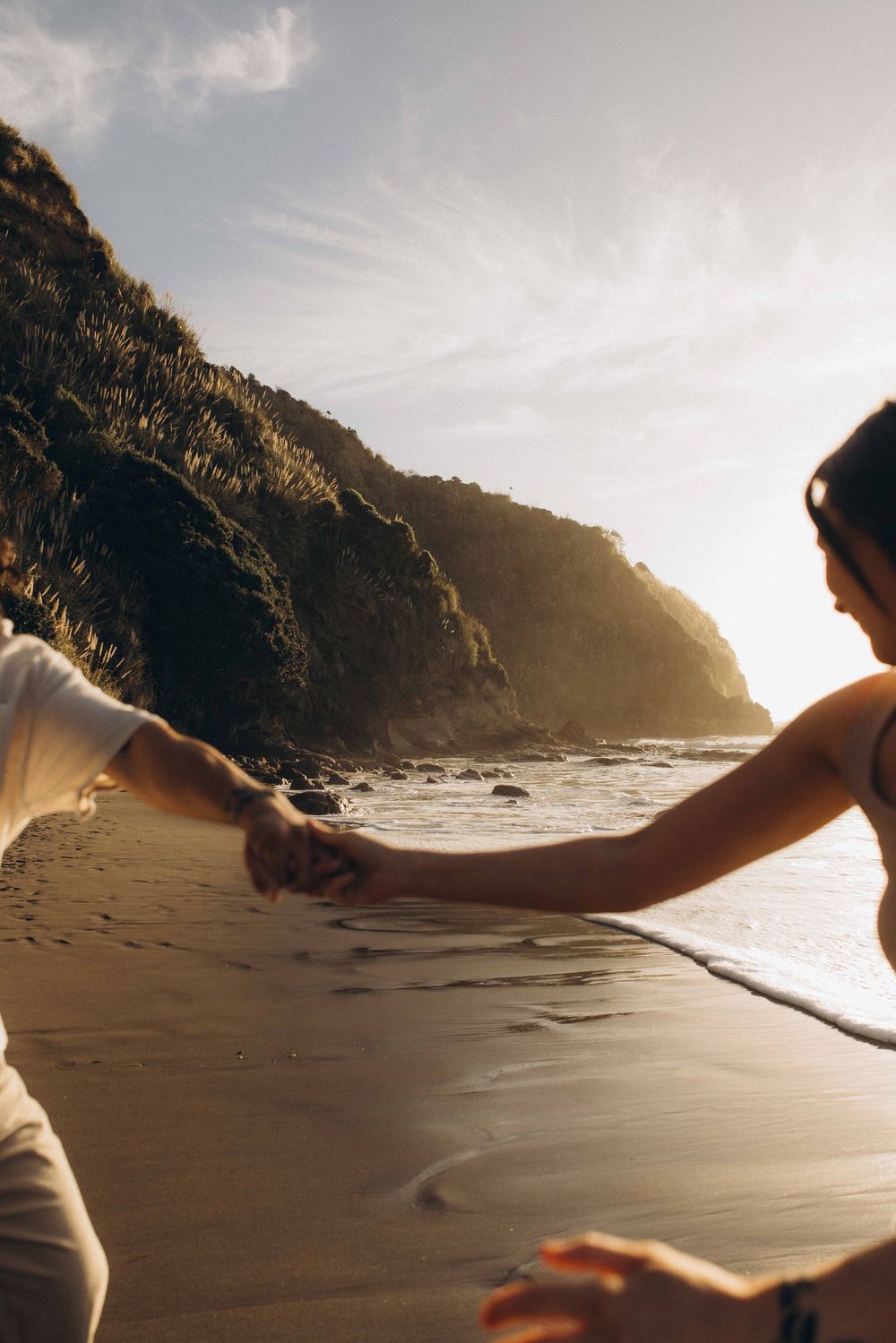 Couple holding hands on a sunlit sandy beach with dramatic cliffs and ocean waves in the background.