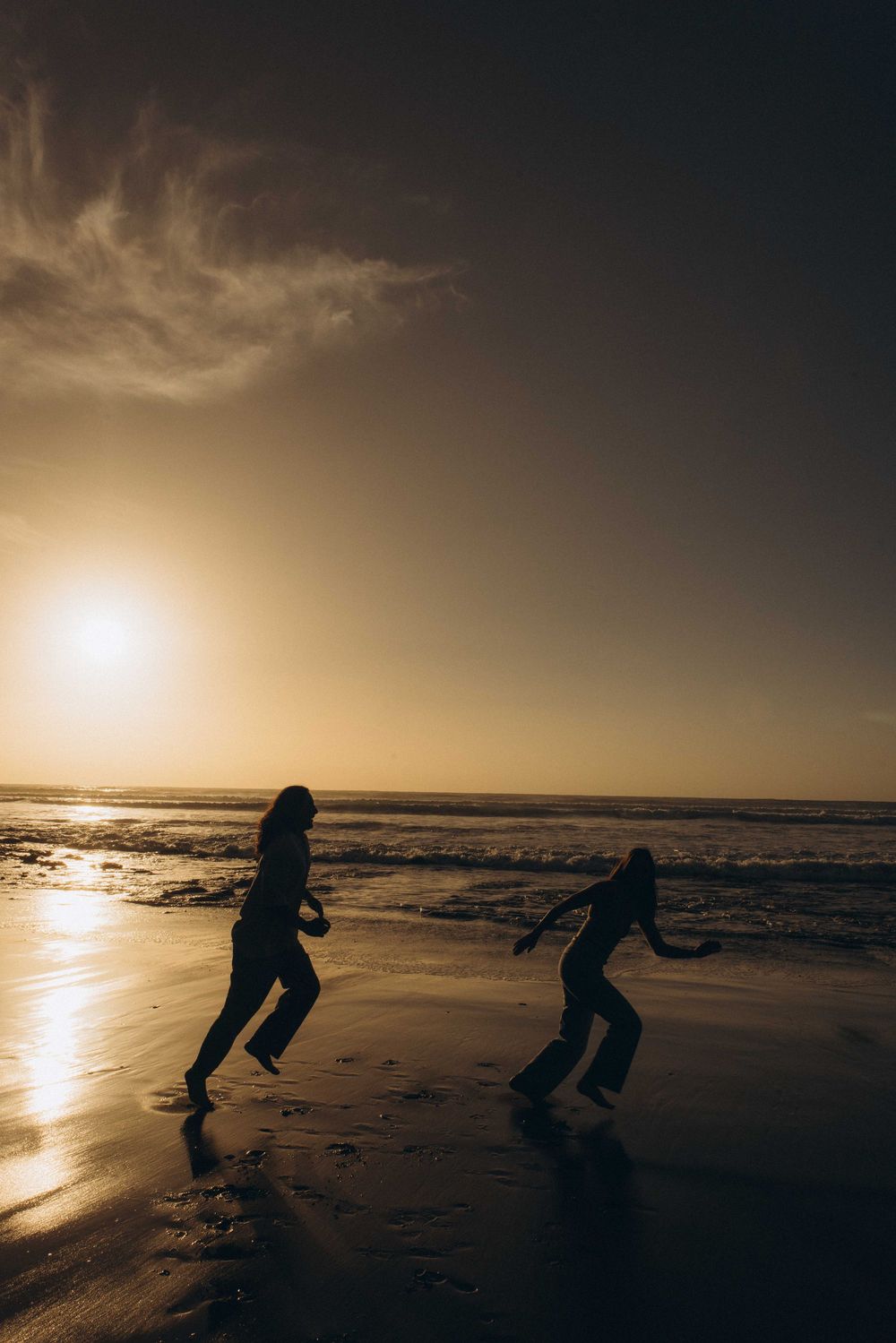 Two silhouettes play on a reflective beach at golden sunset with ocean waves in the background.