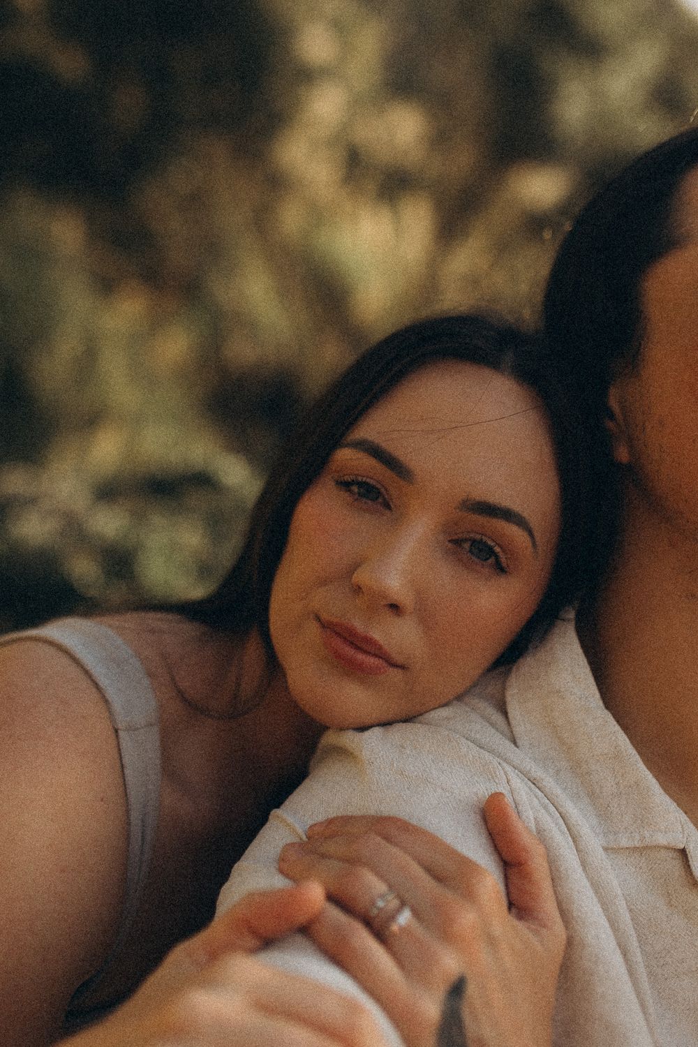 A woman with dark hair rests her head on someone's shoulder, holding hands, with a soft bokeh background.