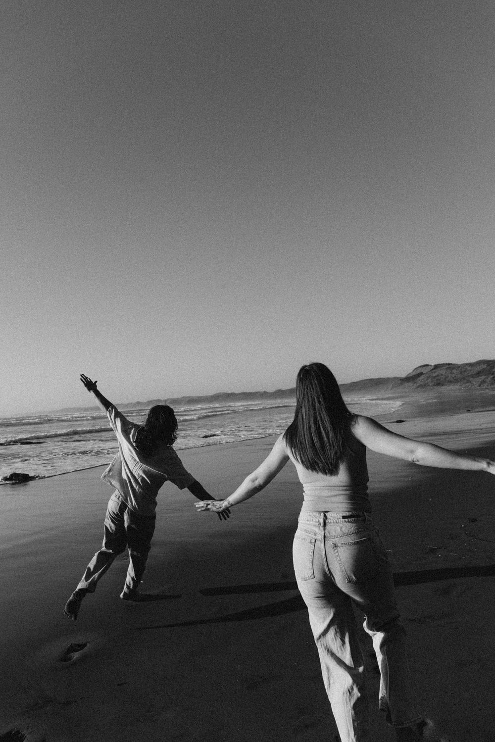 Black and white photo of two people holding hands on a beach, arms outstretched, facing the ocean waves.