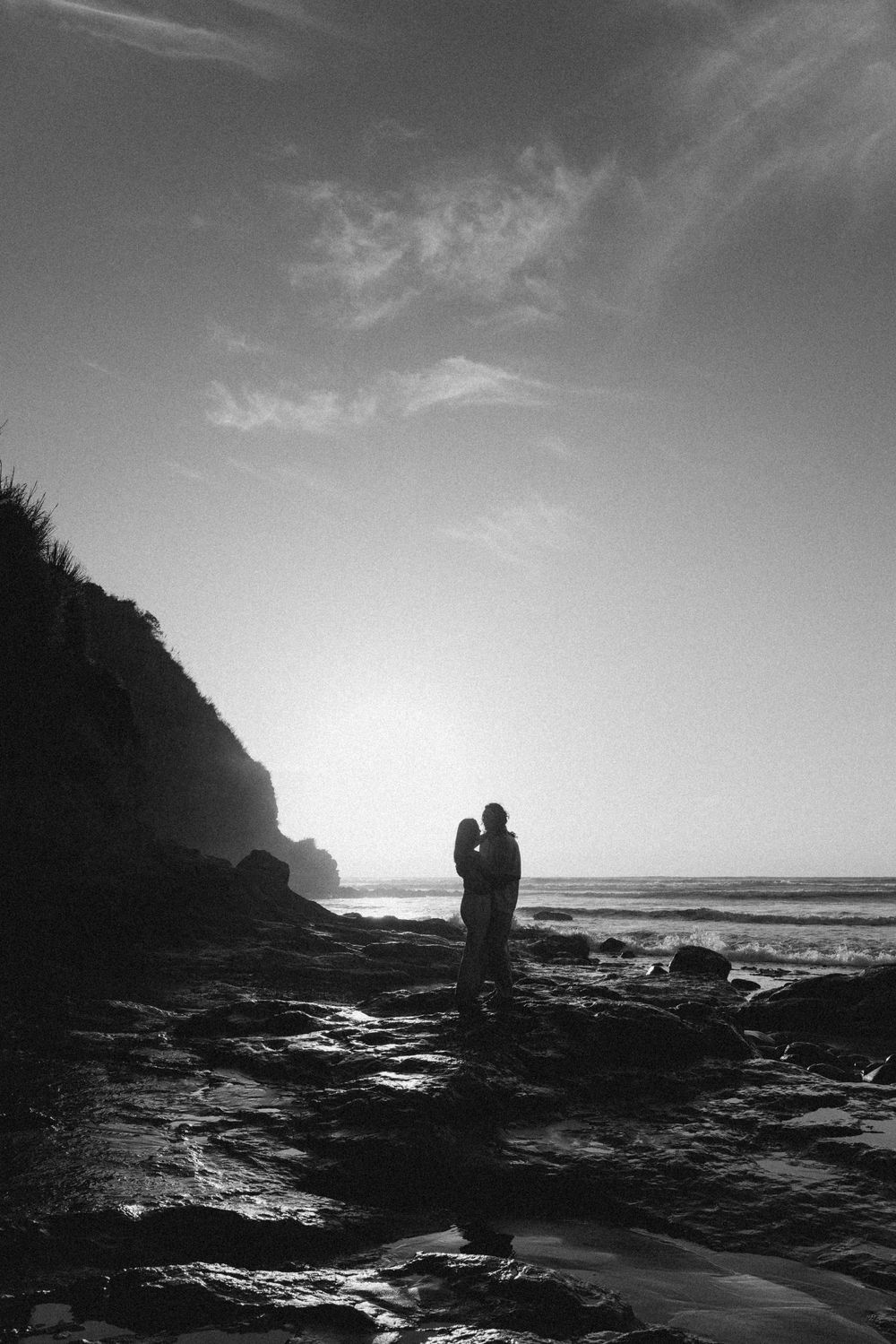 Black and white silhouette of a couple embracing on a rocky beach at sunset with cliffs and ocean waves.