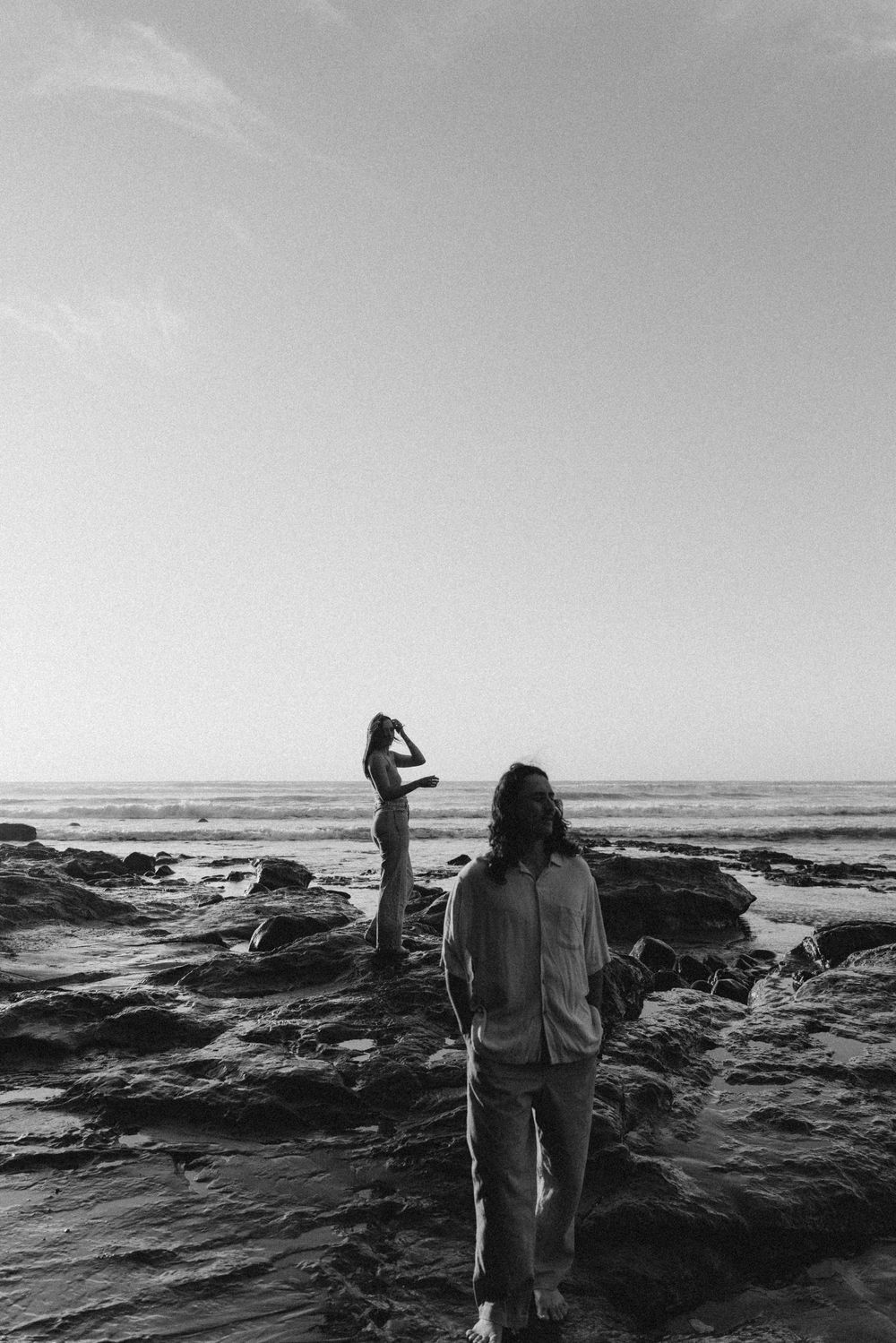 Black and white photo of two people standing on rocky beach shore with ocean waves in the background.