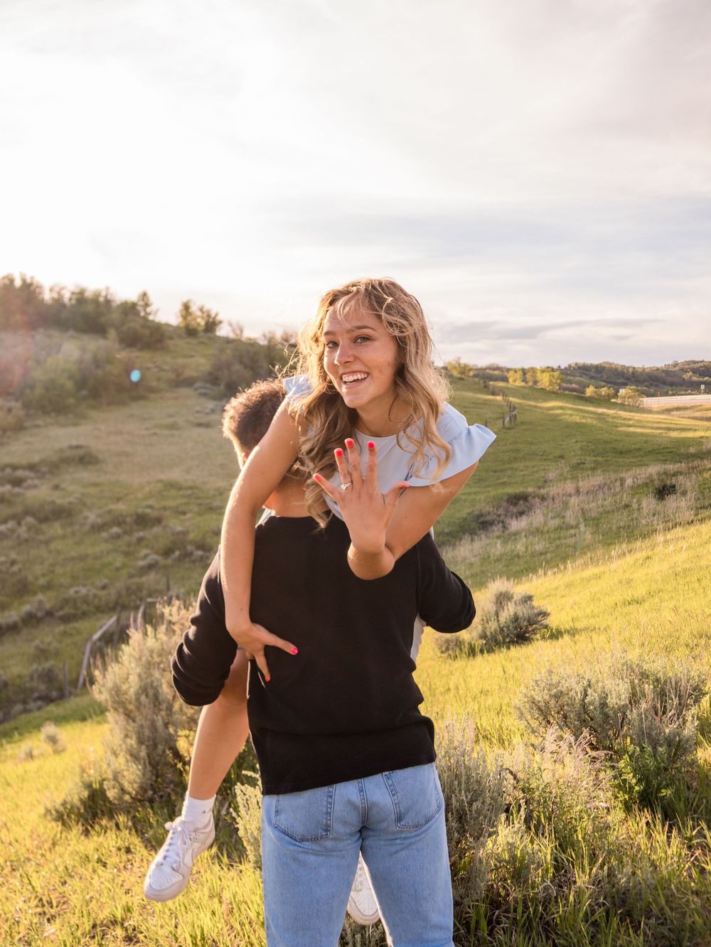 A joyful woman shows off her engagement ring while getting a piggyback ride outdoors on a sunny hillside.