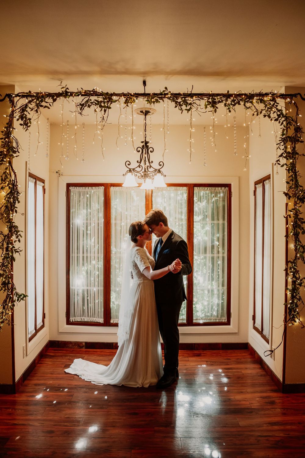 Bride and groom share first dance under fairy lights and floral arch in elegant indoor wedding venue.