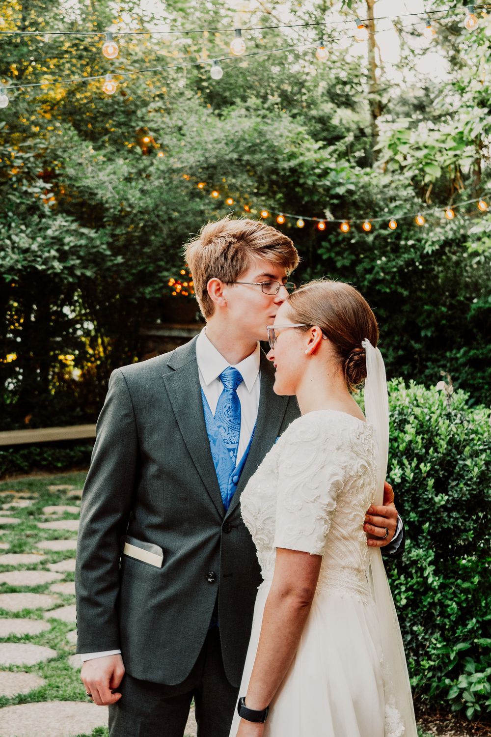 A bride and groom share a romantic kiss in a lush garden setting, with string lights and greenery in the background.