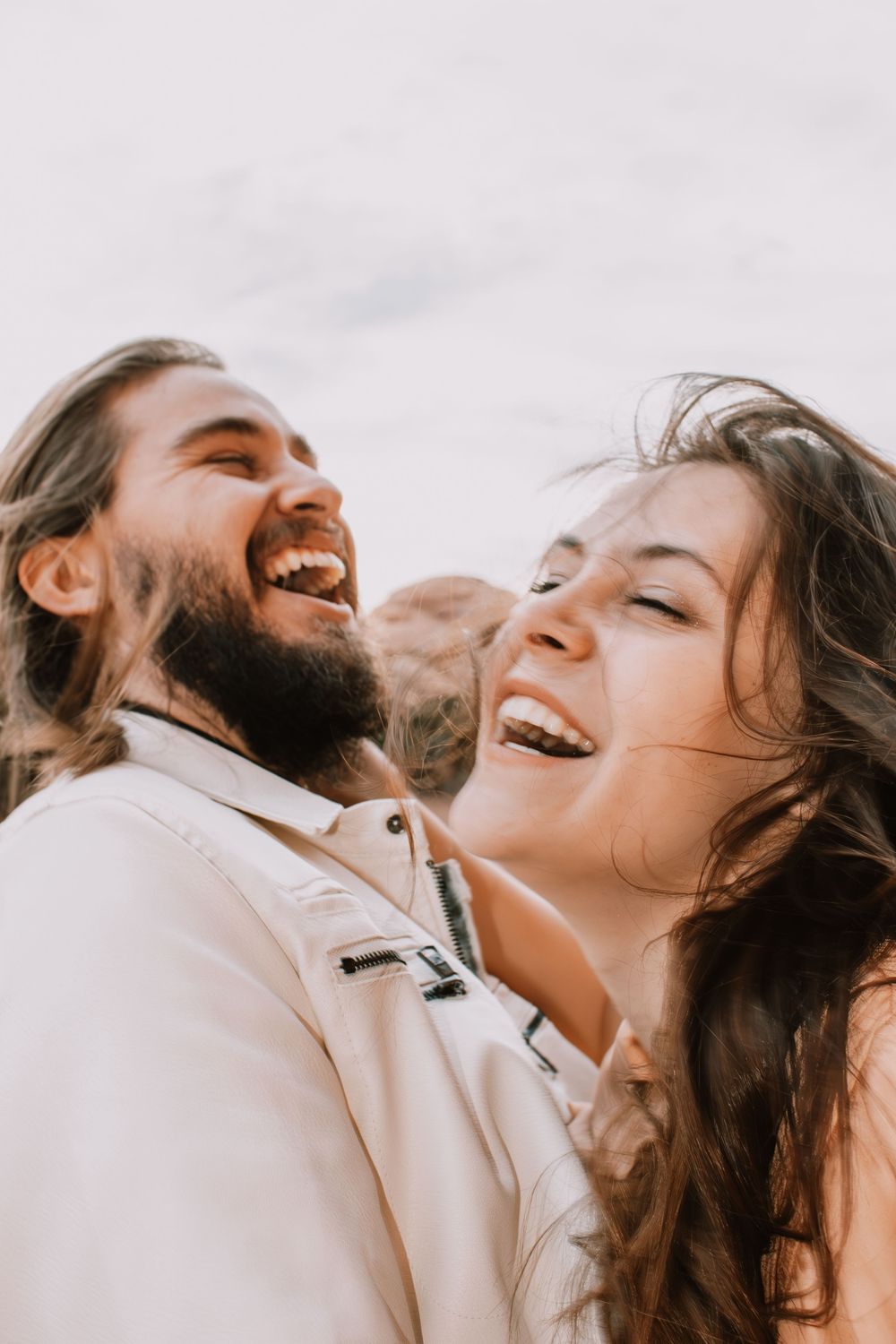 A laughing couple with long hair and beard enjoying a joyful moment together outdoors on a bright day.