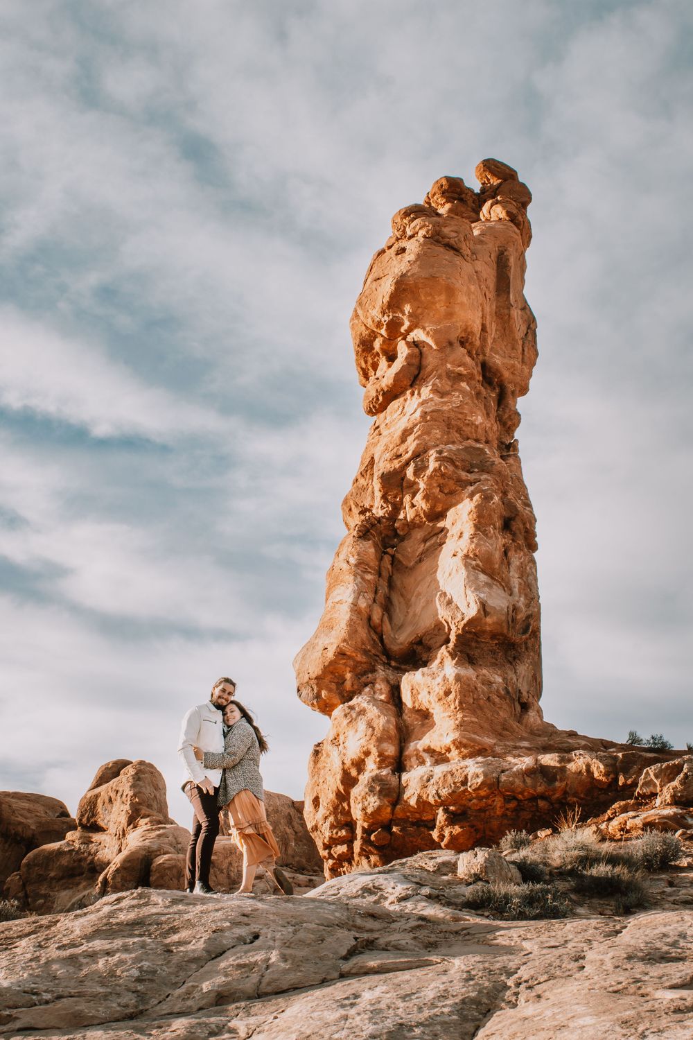 Couple embracing beside a tall red rock formation in a desert landscape under a partly cloudy sky.