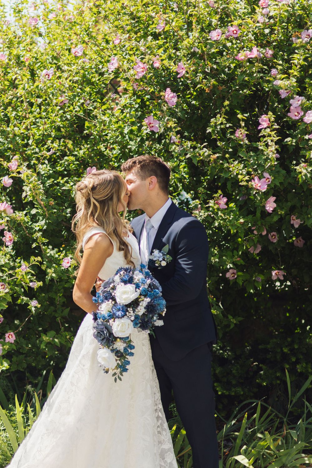 Bride and groom kissing in front of lush green bushes with pink flowers, bride holding blue and white bouquet.
