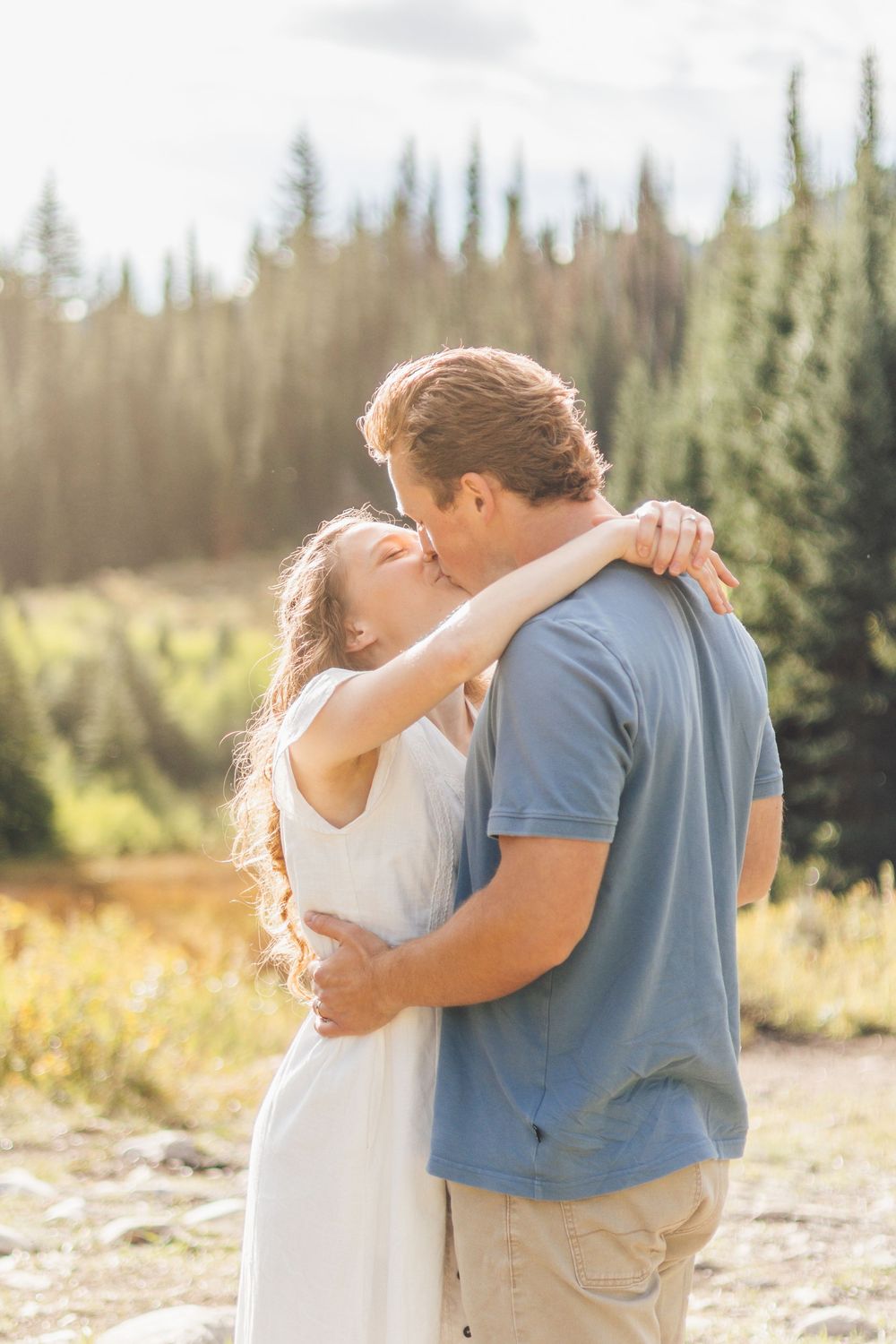 Couple sharing a romantic kiss outdoors with pine trees and golden meadow in background during sunset.