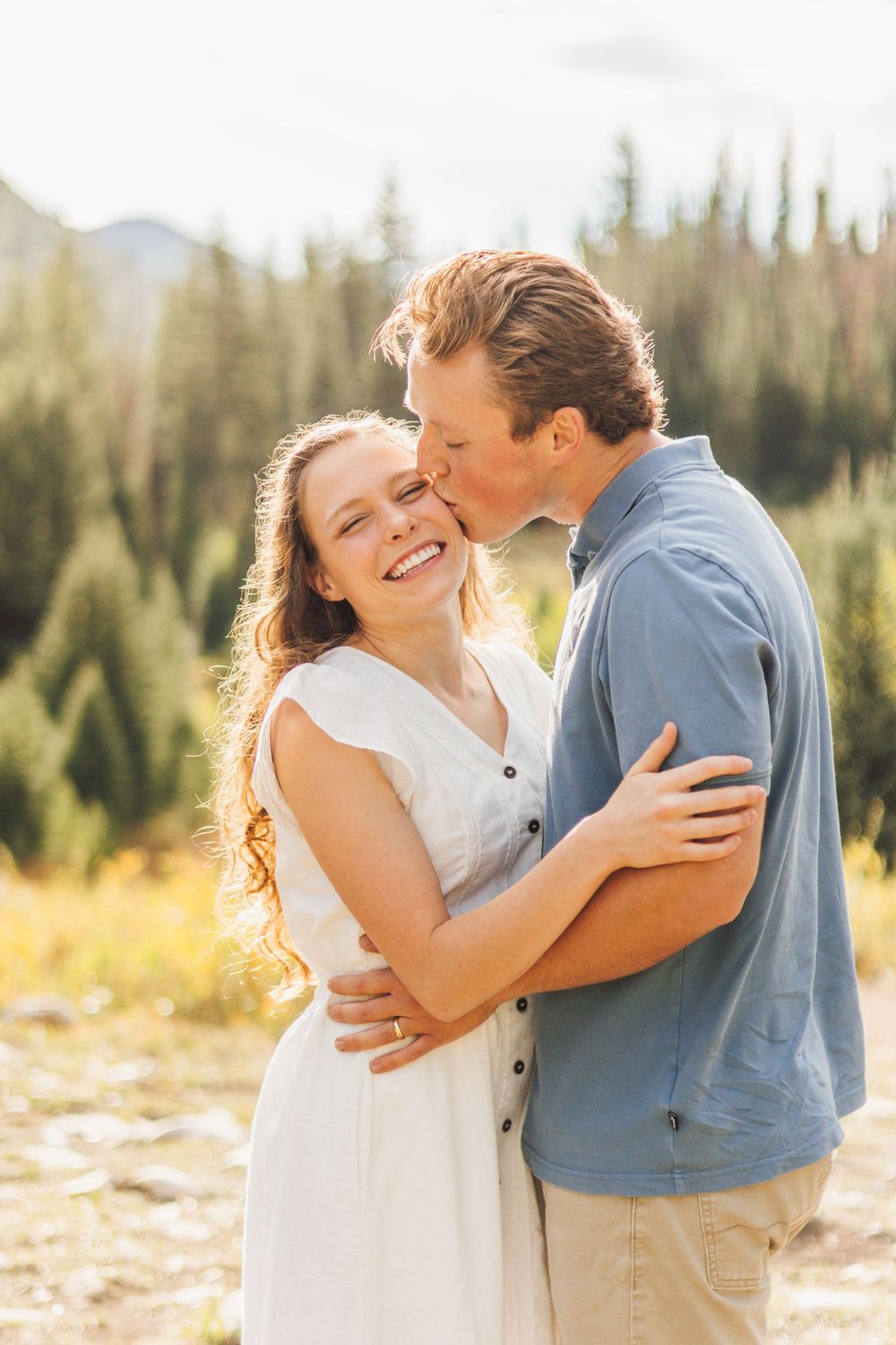 A couple embraces outdoors, woman in white dress smiling as man kisses her cheek, with pine trees in background.
