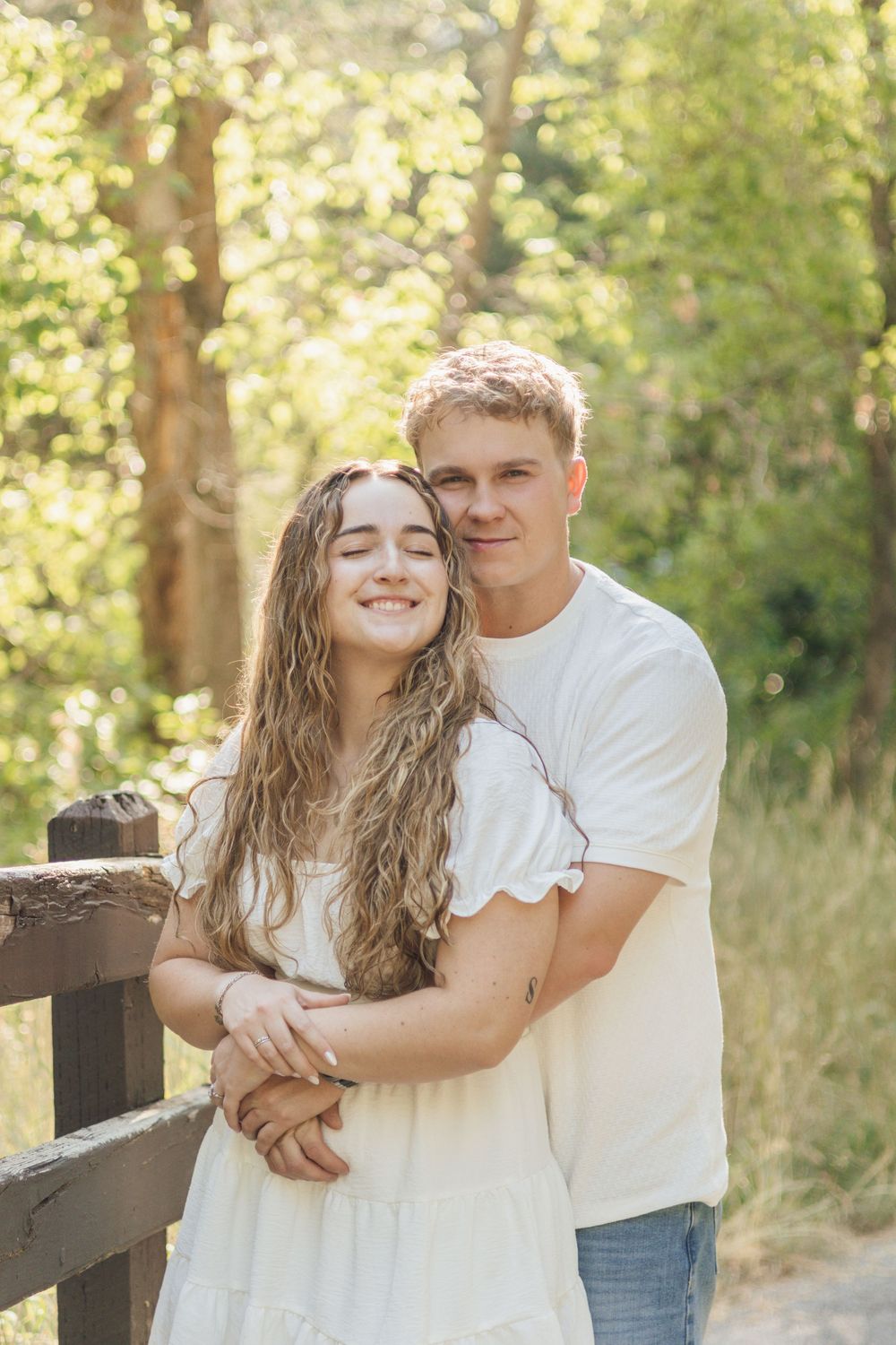 Young couple embracing on a wooden bridge in a sunlit forest, both wearing white, smiling warmly.