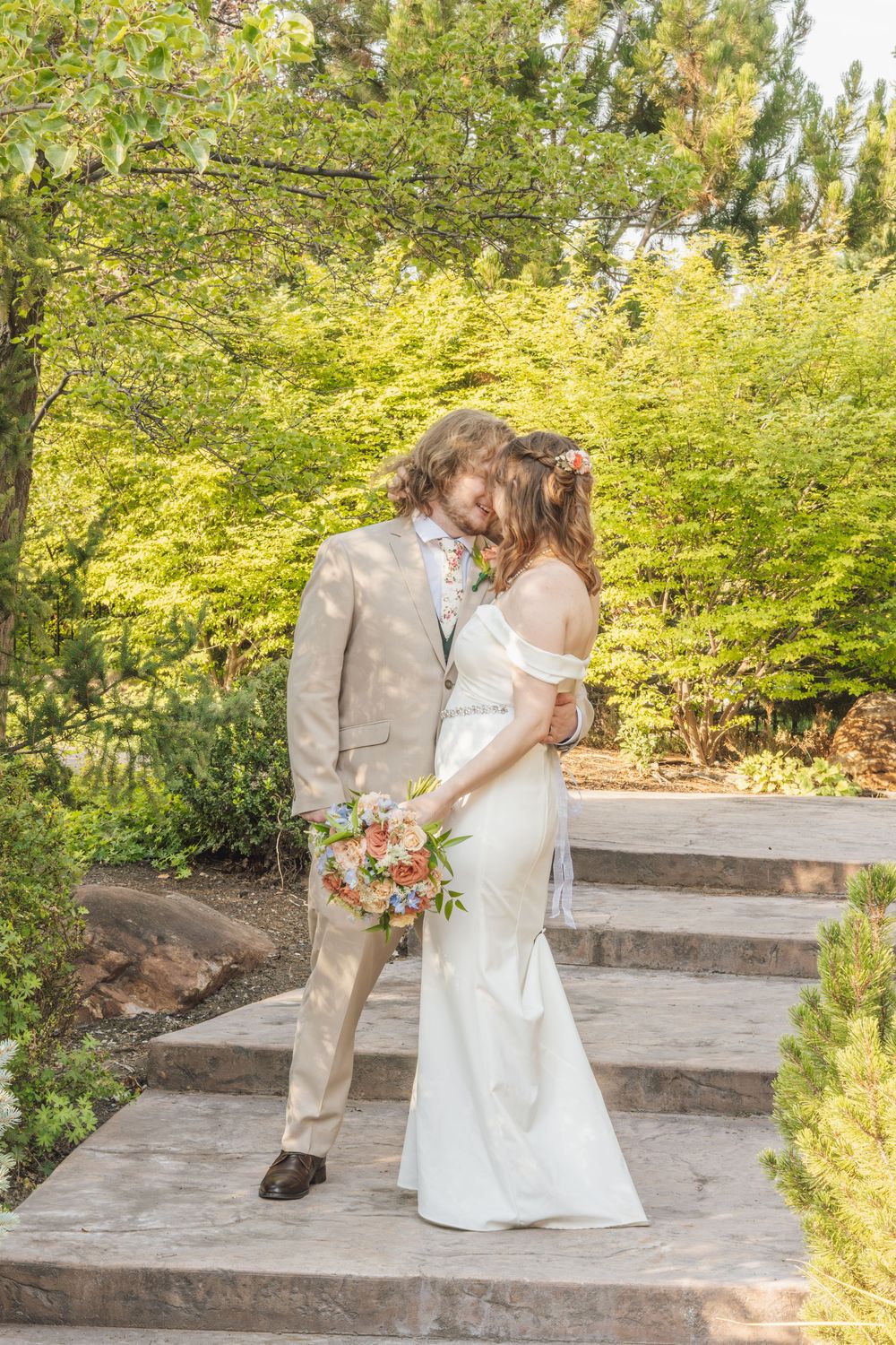 Bride and groom share a romantic kiss outdoors, surrounded by lush green trees, as bride holds a colorful bouquet.