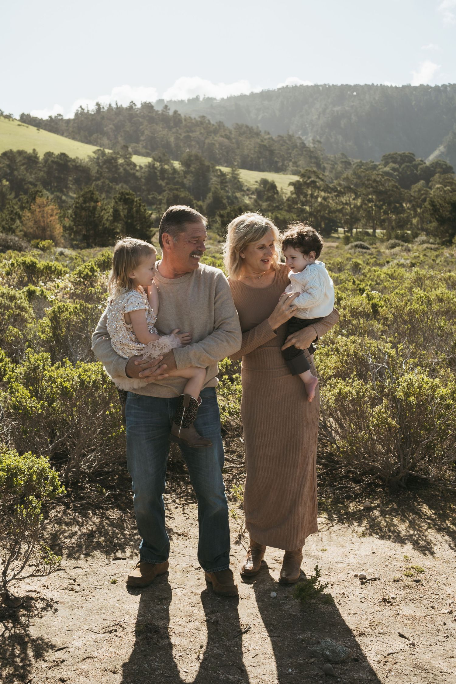 Family of four with two young children posing outdoors in a sunlit green hillside landscape.