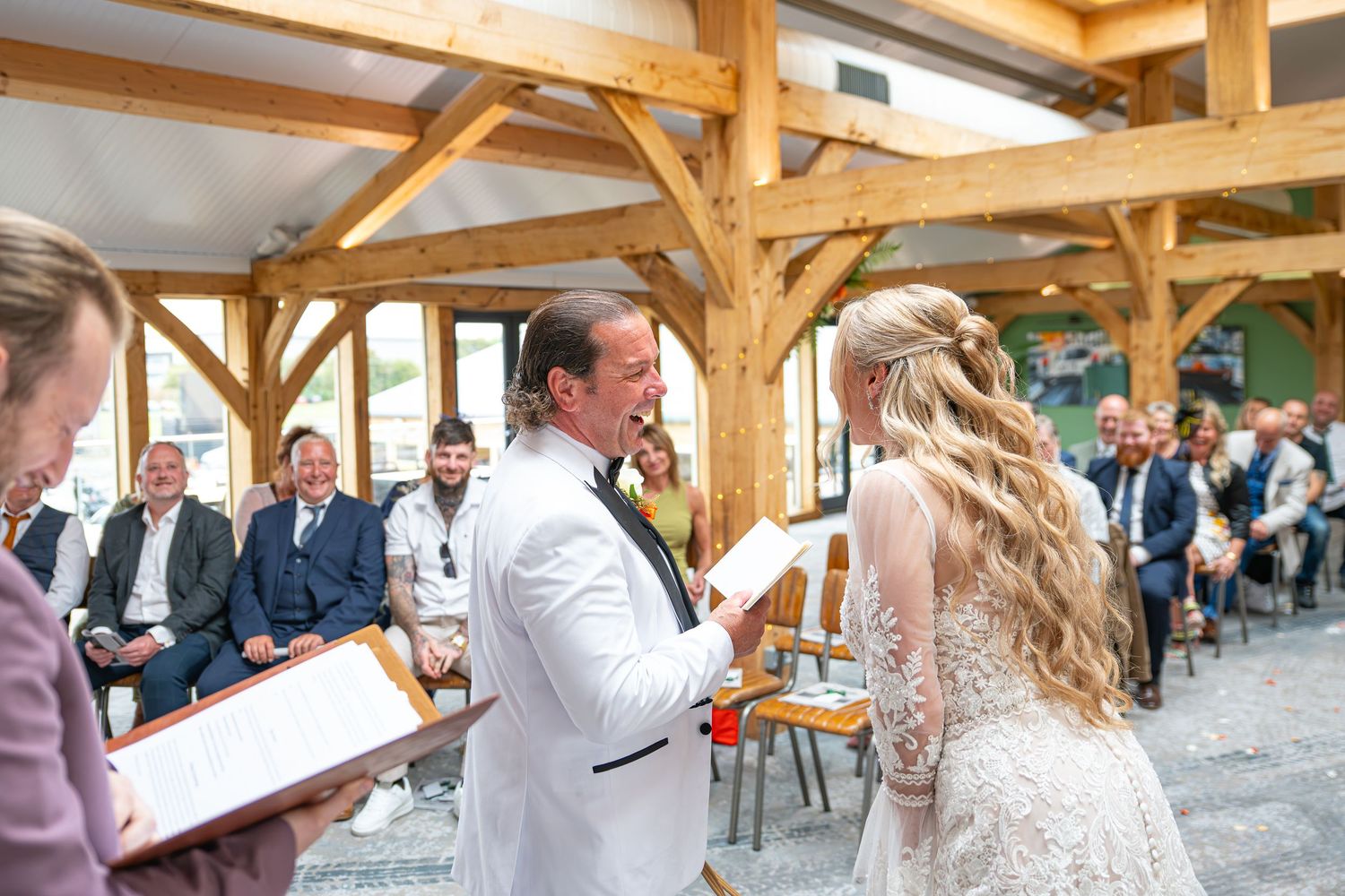Groom in white suit reads vows to bride in lace dress during wedding ceremony with seated guests.Yorkshire, Sheffield, Lincolnshire, UK