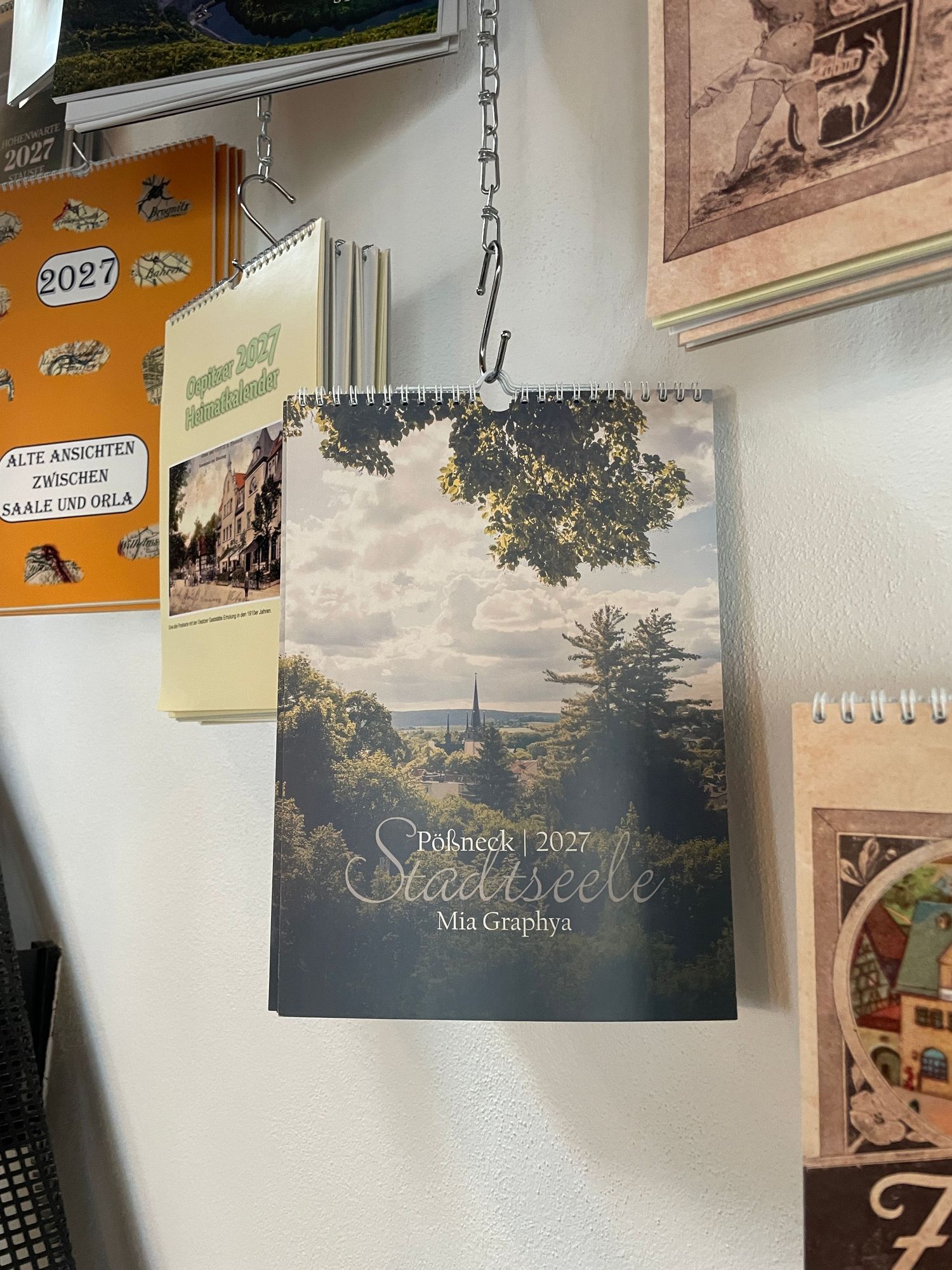 A 2027 Pößneck Stadtseele calendar by Mia Graphya displayed on a wall among other calendars.
