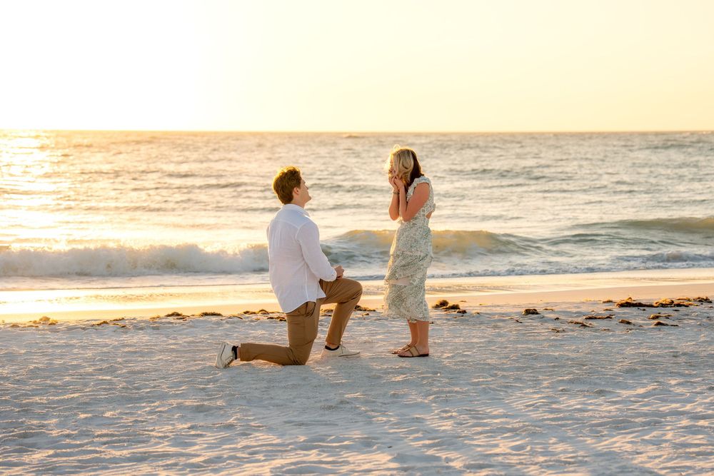Man proposing on one knee to surprised woman on sandy beach at golden sunset.