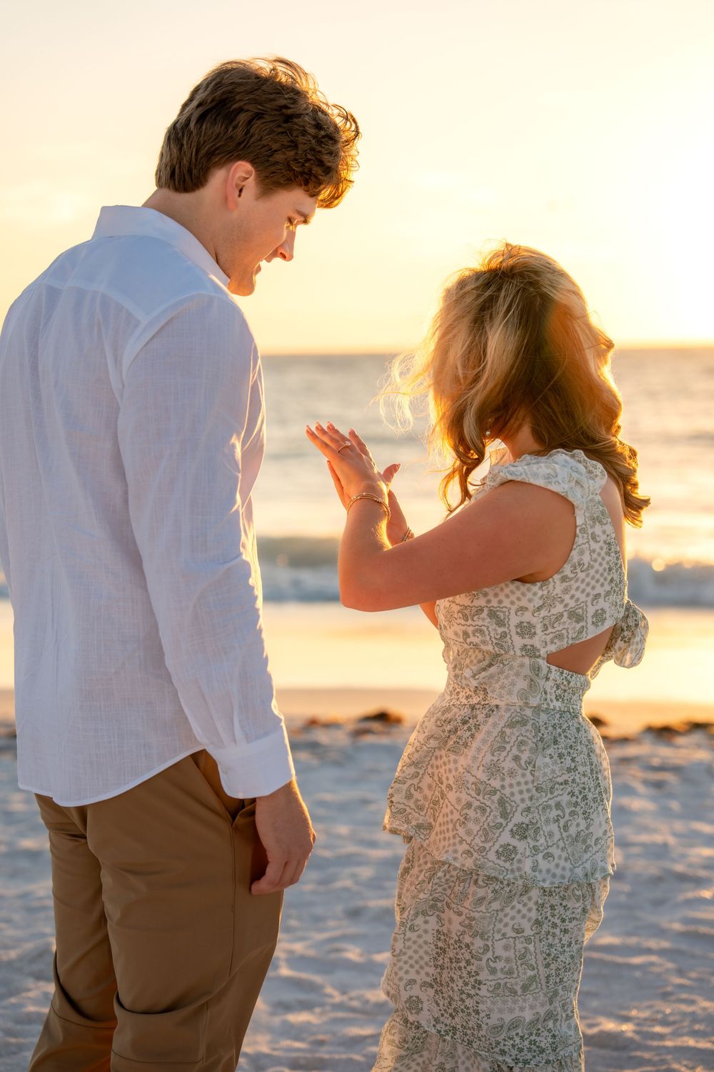 Newly engaged couple admiring engagement ring on beach at golden hour sunset.