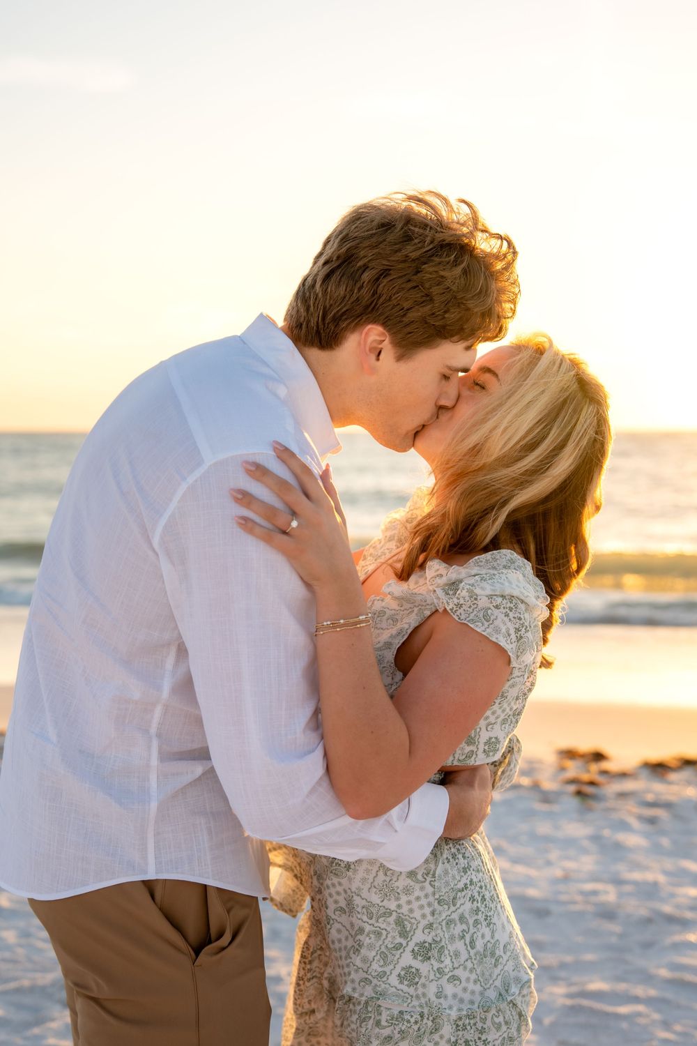 Newly engaged couple sharing passionate kiss on beach bathed in warm golden sunset light.