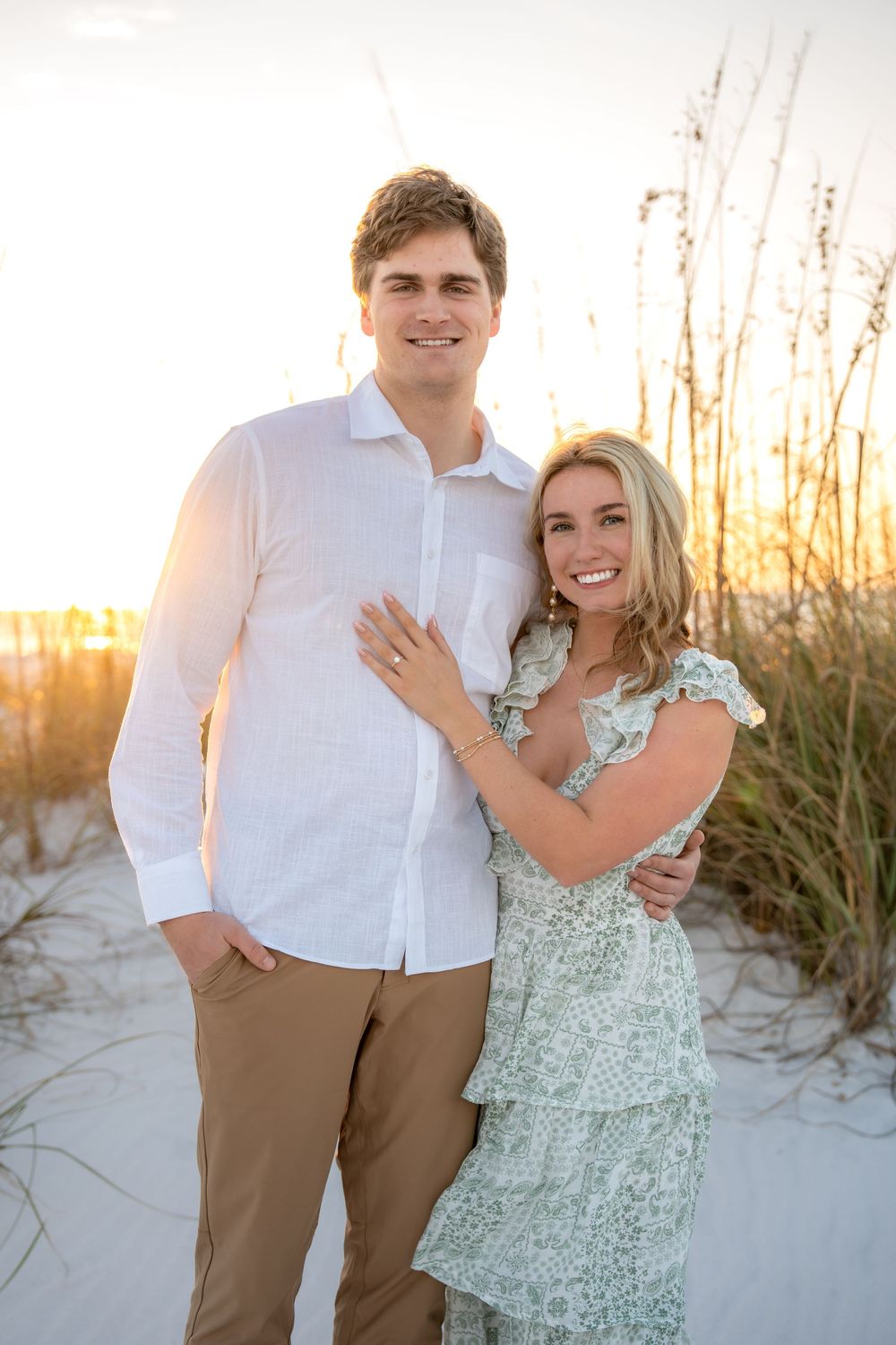 Smiling newly engaged couple posing in front of sea oats on white sand beach at sunset.
