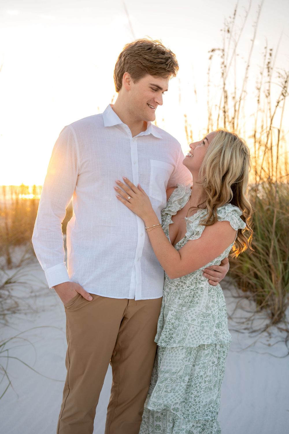 Couple sharing a romantic moment at sunset on a sandy beach, woman in floral dress embracing man in white linen shirt.