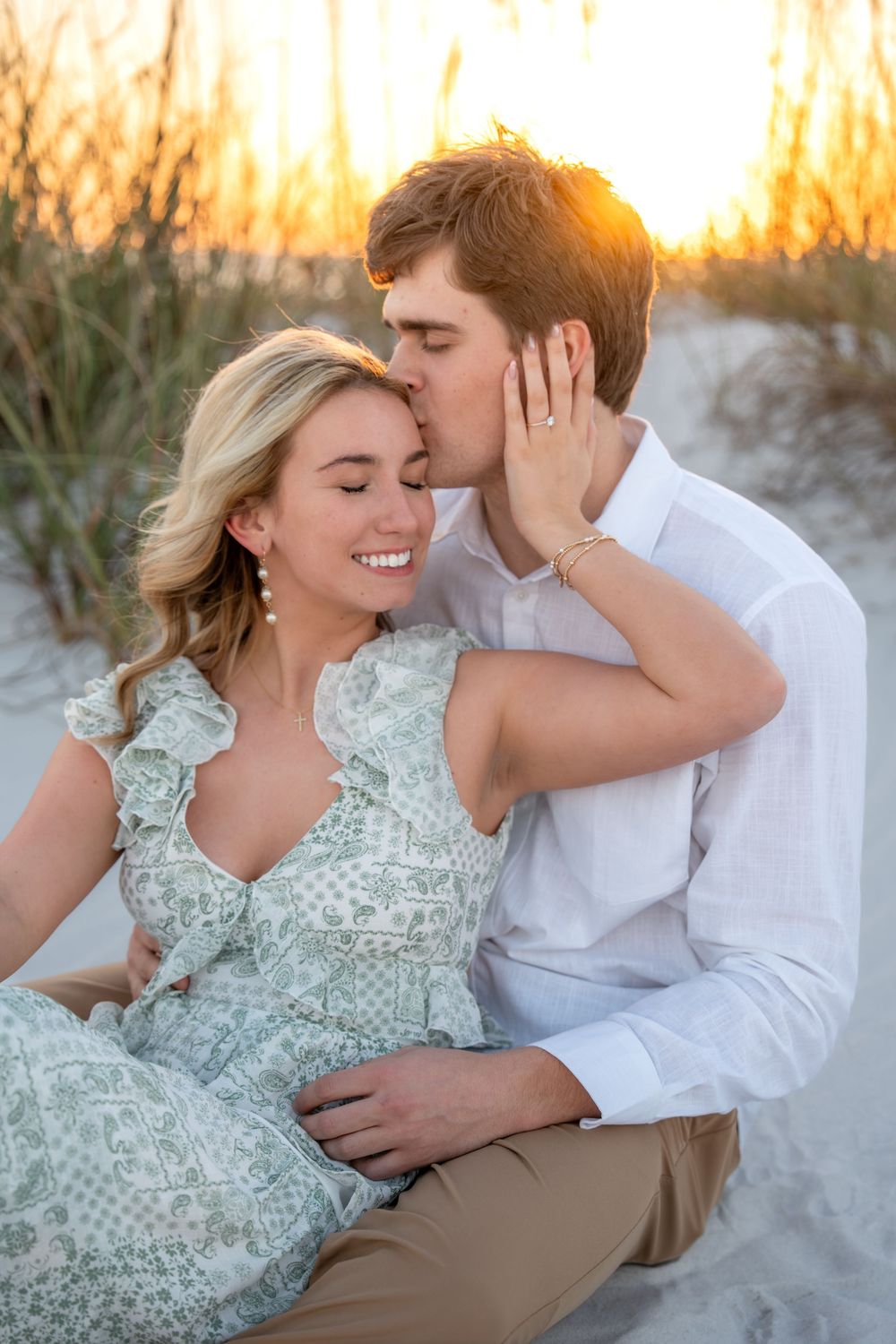 Man tenderly kissing woman's cheek at golden hour beach sunset, woman smiling in sage green floral dress.