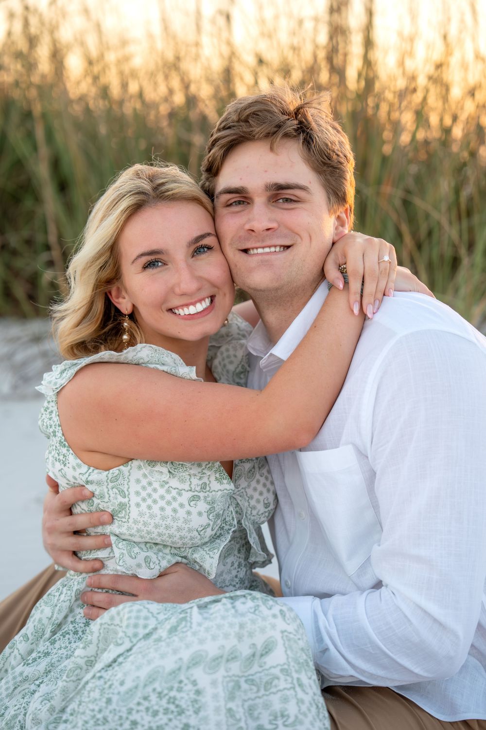 Happy engaged couple embracing and smiling on beach at sunset, woman in green floral dress hugging man in white shirt.