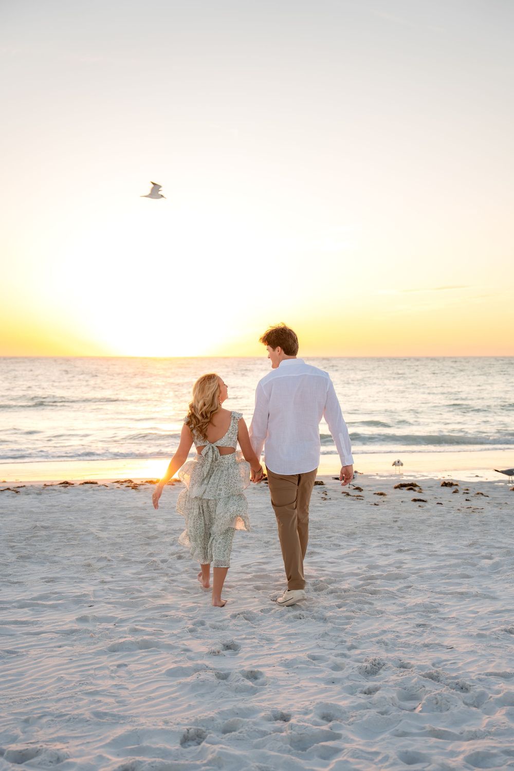 Couple holding hands walking toward ocean at stunning beach sunset, woman in floral dress and man in white shirt.