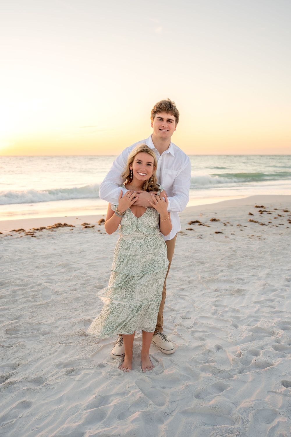 Man embracing woman from behind on sandy beach at sunset, couple smiling in coordinated beach engagement outfits.