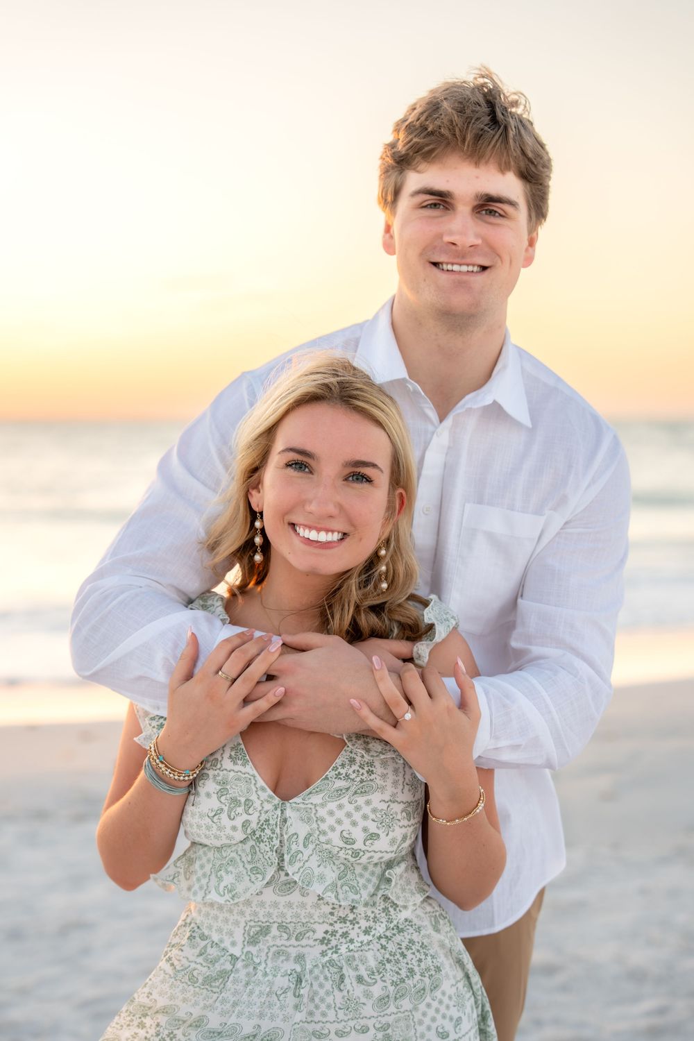 Engaged couple posing at beach sunset, man in white shirt embracing woman in sage green floral dress from behind.