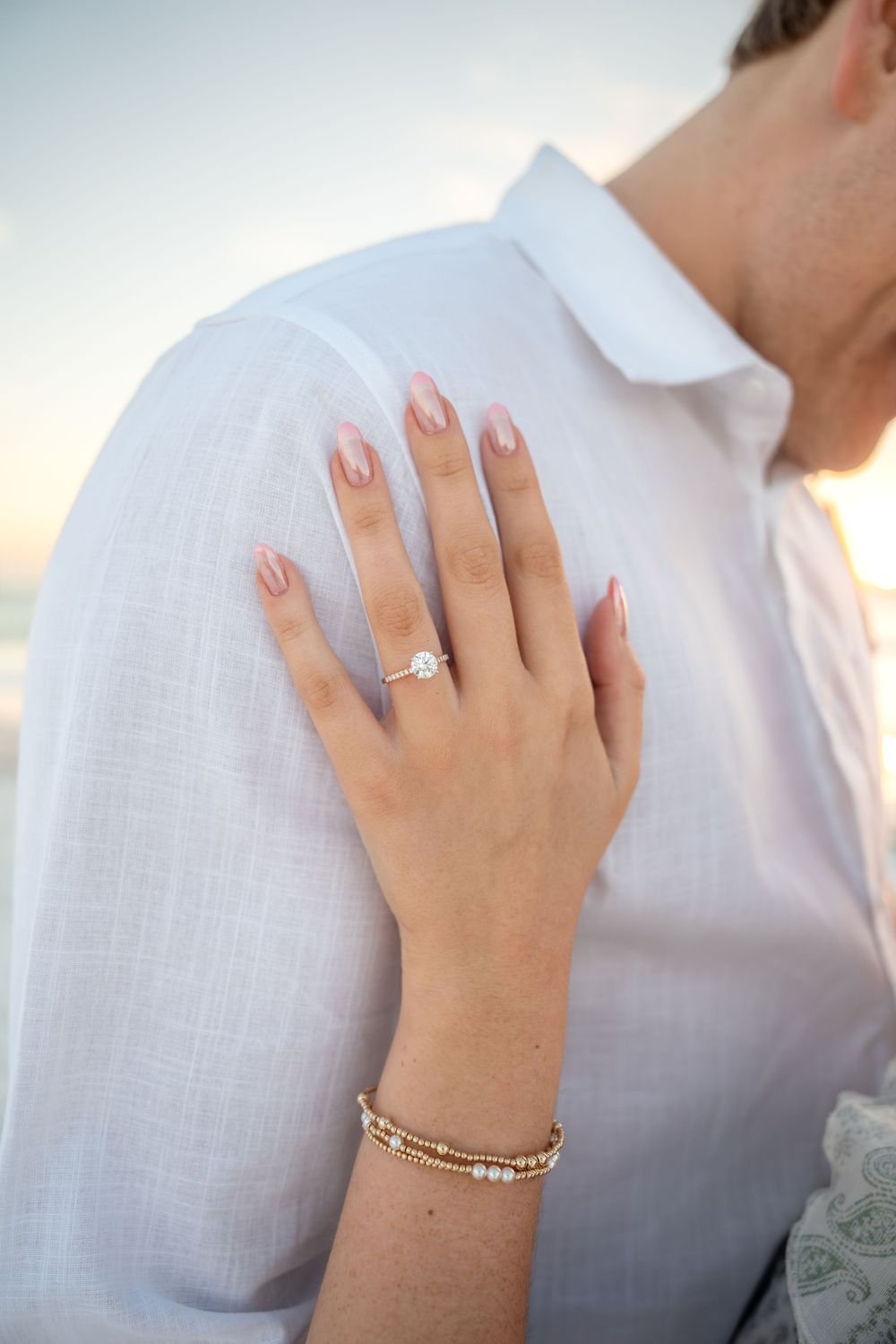 Close-up of woman's hand displaying diamond engagement ring on man's white linen shirt shoulder at beach.