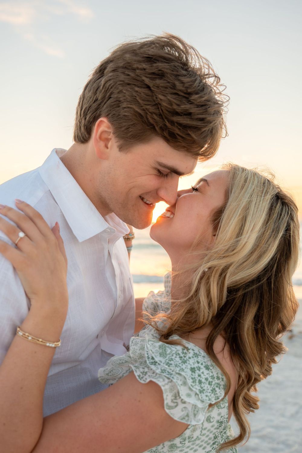 Engaged couple sharing a kiss at golden beach sunset, woman in floral dress with diamond ring visible on hand.