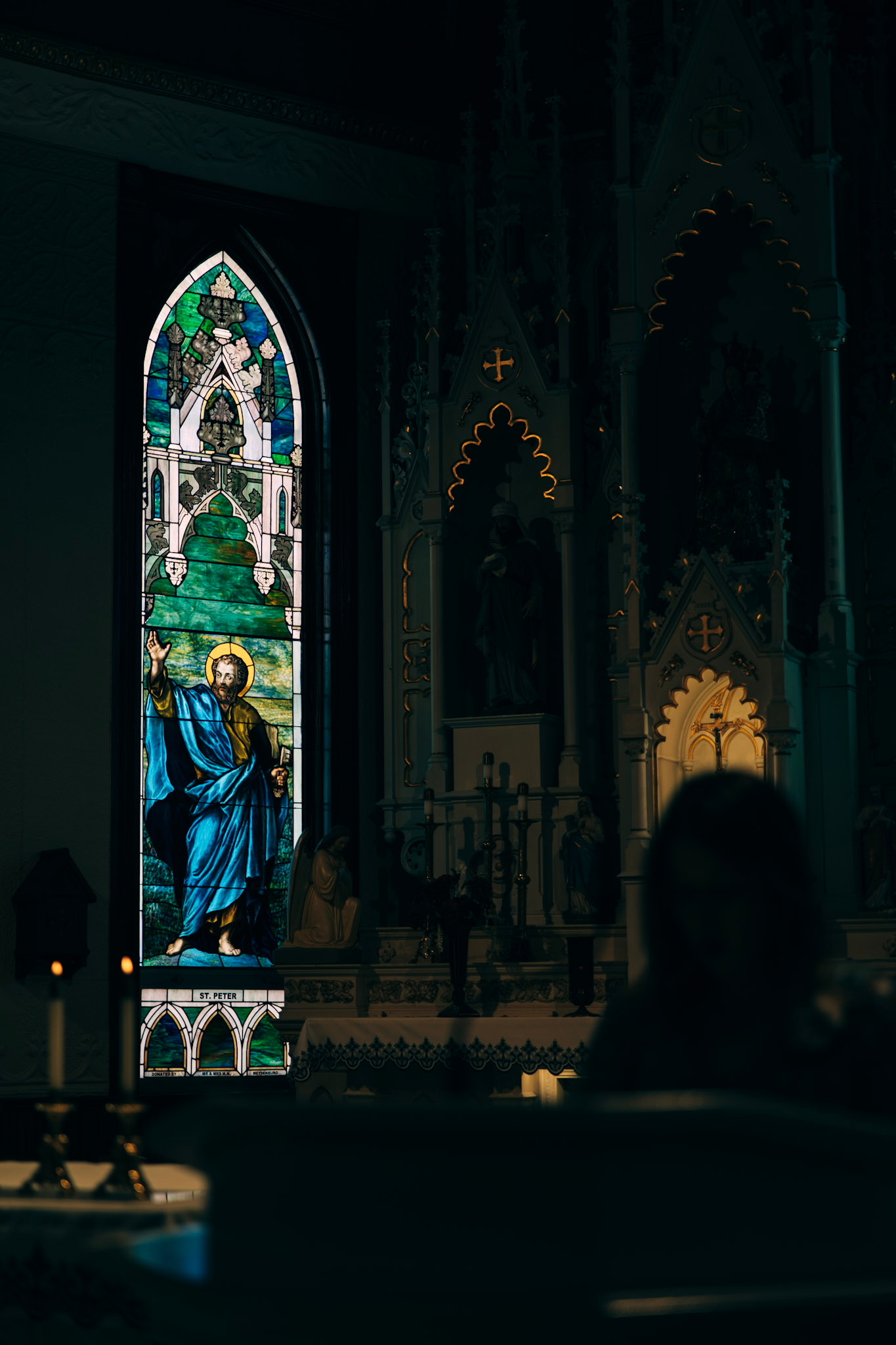 A worshipper sits in a dimly lit church, illuminated by a vibrant stained glass window depicting a saint in blue robes.